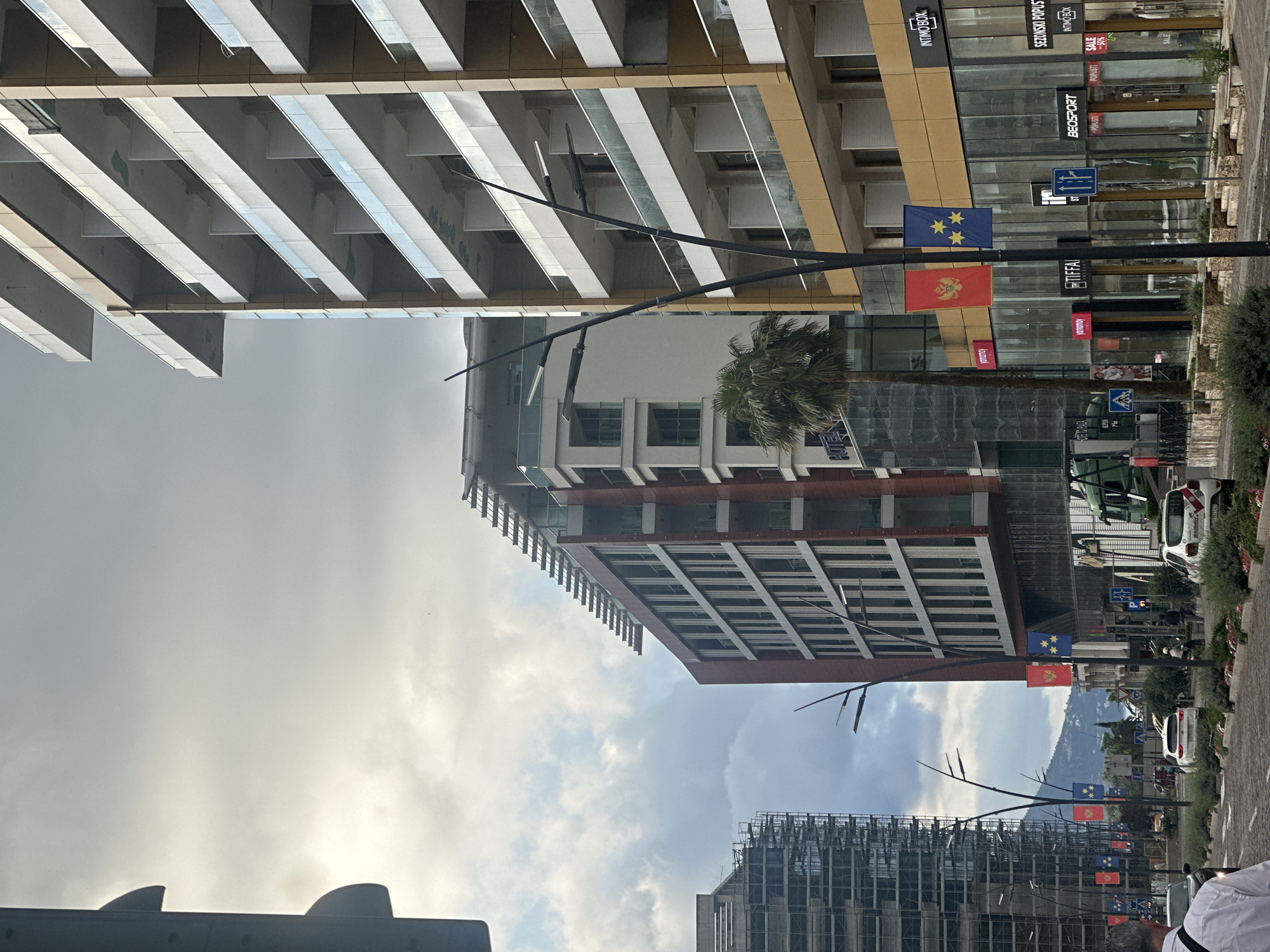 The image, taken in Budva, Montenegro, shows a street scene with several modern buildings and flags. On the right side of the image, there are glass-fronted storefronts with various brand names displayed, including "INTIMBOX," "SEZONSKI POPUS," "BEOSPORT," and "TIFA." The flags of Montenegro and what seems to be the flag of Budva are attached to a light pole in front of this building. The architecture is predominantly modern, with buildings featuring glass and concrete elements. Above and behind the stores, there's a taller, multi-story structure with a red and white facade. In the background, a partially constructed building is visible. The sky is overcast with thick clouds. A palm tree is standing beside the glass-front building. Vehicles, including what appears to be a police car, are parked along the street. The street itself is lined with greenery and street furniture, and blurred figures suggest pedestrian activity.