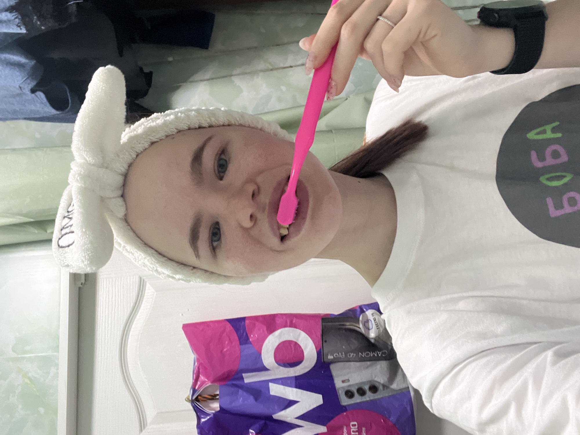 A person with a white headband and a white t-shirt with "BOBA" written on it is brushing their teeth with a pink toothbrush. The person is wearing a black smartwatch. A purple and pink package with the word "WOW" and "CAMON 40 Pro" is visible. A white door with moldings is also visible in the background. The location is implied to be in Chaikovsky, Russia.