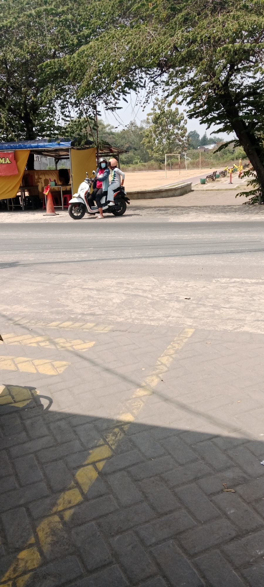 In Maguwoharjo, Indonesia, on a sunny day, two individuals are seated on a white scooter on a paved road. The person in the front is wearing a blue shirt, black pants, and a white face mask, with a dark helmet. The person behind them wears a gray long-sleeved shirt, white pants, and a light orange helmet. They are positioned near a yellow awning-covered stall with orange cones. Beyond the road, a sandy field with a white goalpost and benches suggests a recreational area or sports field. Trees with green foliage frame the scene. The foreground shows a patterned paved surface with yellow markings, possibly indicating a parking area or crosswalk.