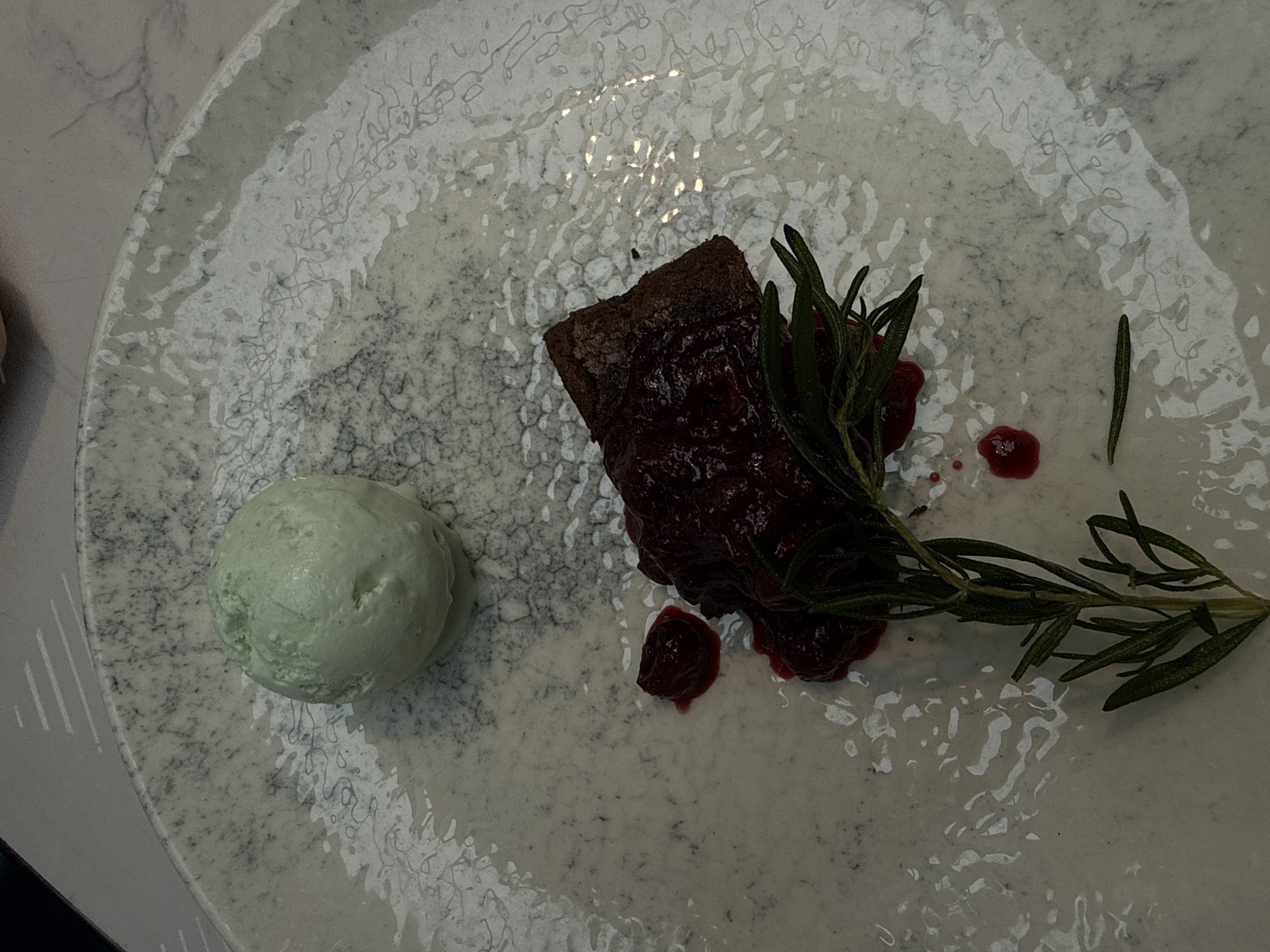 The image displays a dessert presented on a light-colored, irregularly patterned plate with grey and black markings. The specific geographical location in Izhevsk, Russia, cannot be determined from the visual information provided.
In the upper-left section of the plate, there is a spherical scoop of light green ice cream. Towards the lower-center, a rectangular piece of dark brown cake or brownie is visible, topped with a glossy, dark red berry compote or sauce. A fresh sprig of green rosemary lies diagonally across the cake and extends towards the lower-left. Several distinct drops and smears of the dark red sauce are scattered on the plate surface around the cake and rosemary. A single detached rosemary leaf is situated near the bottom-left edge of the plate. The plate surface exhibits reflections of overhead lighting. No individuals are present. The background, partially visible, suggests a light-colored, possibly textured table or countertop surface.