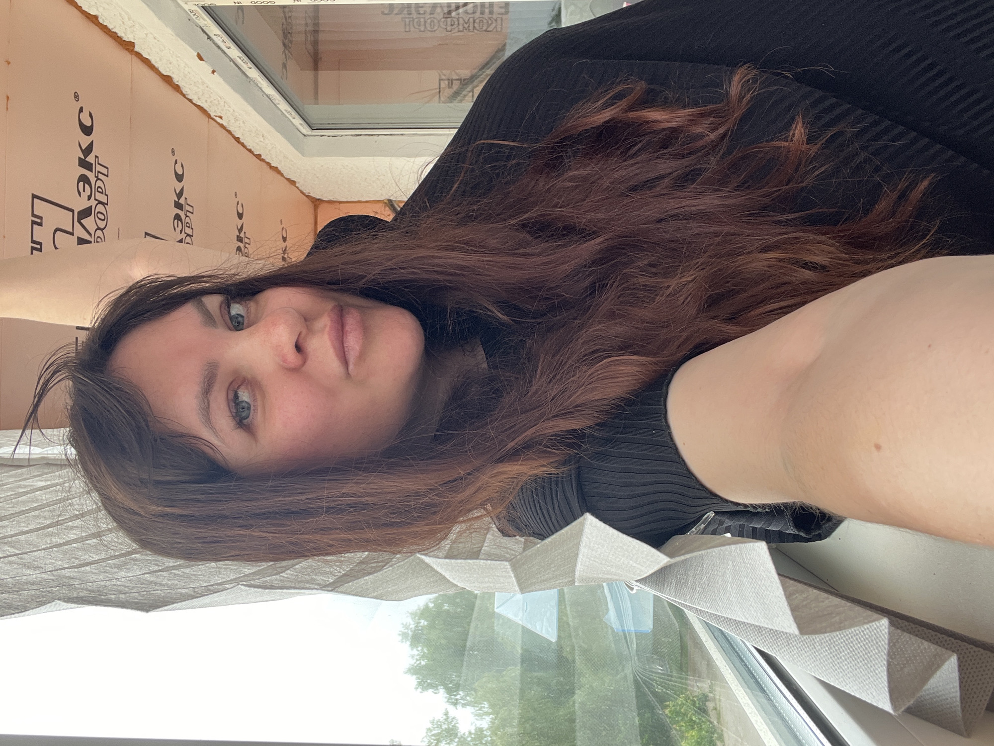 This is a portrait-oriented selfie featuring a woman looking directly at the camera with a neutral to slightly pensive expression. She has fair skin, blue-green eyes, and long, wavy, dark reddish-brown hair, which falls loosely around her shoulders. She is wearing a black, ribbed long-sleeved top.
The setting appears to be indoors, likely in a room undergoing some renovation or construction, as indicated by the background. To the woman's right, a modern white UPVC window frame is visible, partially covered by light grey or beige pleated blinds. Beyond the window, green foliage from trees suggests an outdoor view and clear daytime conditions.
To her left and above her, an orange-pink rigid foam insulation board is exposed, indicating unfinished work around the window. This board has black Cyrillic text printed on it. Clearly visible are parts of the words "ПЕНОПЛЭКС КОМФОРТ" (a specific brand and type of insulation) and "ЭКС" (likely short for "ПЕНОПЛЭКС"). Some reflective text on the window glass, appearing inverted, includes "GOOD IN". The presence of Cyrillic text strongly indicates a location in a Russian-speaking region, aligning with Noviy, Russia. The natural light visible through the window suggests it is daytime with clear or partly cloudy weather. The woman's direct gaze suggests she is taking a self-portrait.