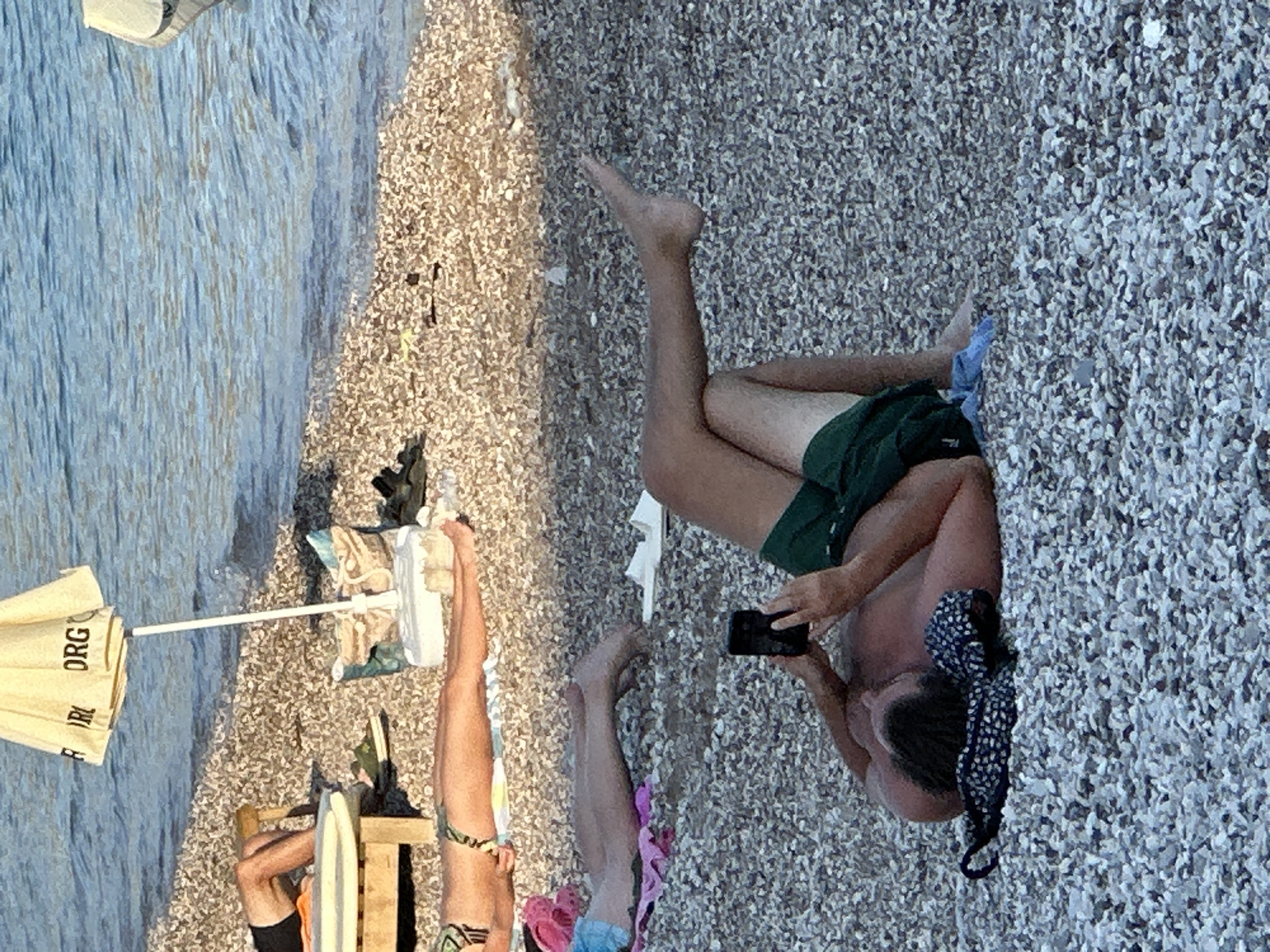 A man is lying on a pebble beach in Budva, Montenegro, holding a black smartphone in his hands. He is wearing green shorts and is shirtless. Next to him on the ground is a patterned bikini top. In the background, other beachgoers are visible, some lying on towels and sun loungers. The sea is to the right of the frame, with gentle waves lapping the shore. An umbrella with the text "ORG" is also visible.