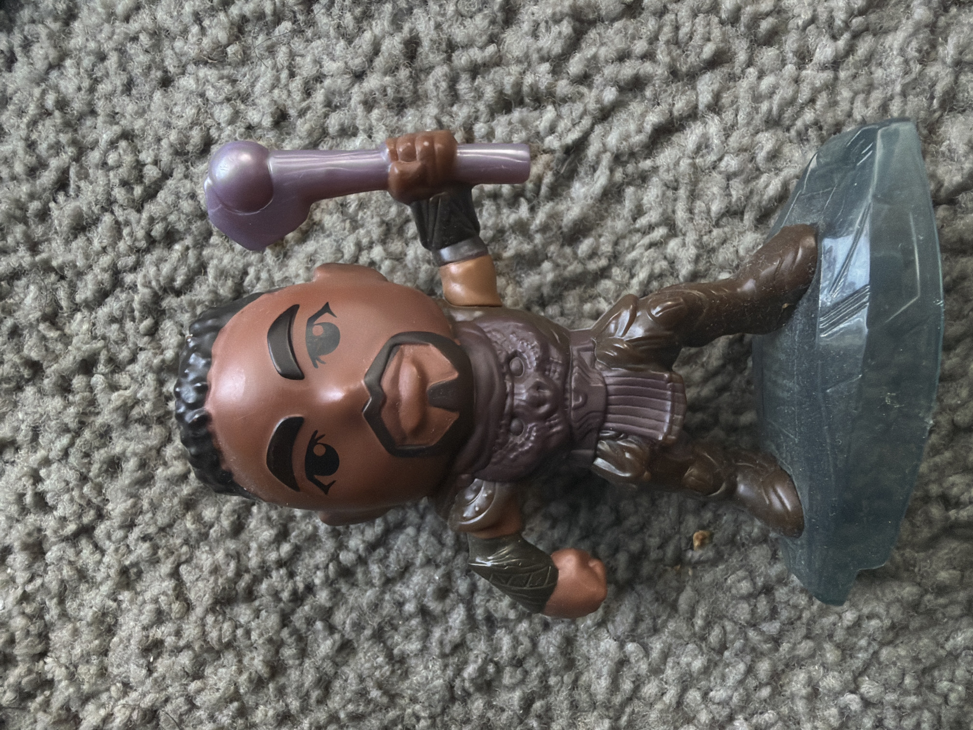 A detailed, full-shot, eye-level, studio photograph shows a toy figurine of a male character standing on a dark gray, rocky base against a muted gray carpet background. The figurine, which appears to be made of plastic, depicts a dark-skinned male with a short black hairstyle and a goatee. He is wearing a brown and purple textured costume that includes armor on his chest and arms, and brown boots. The character is holding a purple club-like object in his right hand. The lighting is bright and even, illuminating the figurine and its details clearly. The carpet background has a high pile texture, with individual fibers clearly visible. The photograph does not depict any specific location within the city of Kennewick, United States of America, as it is a studio shot of a toy.