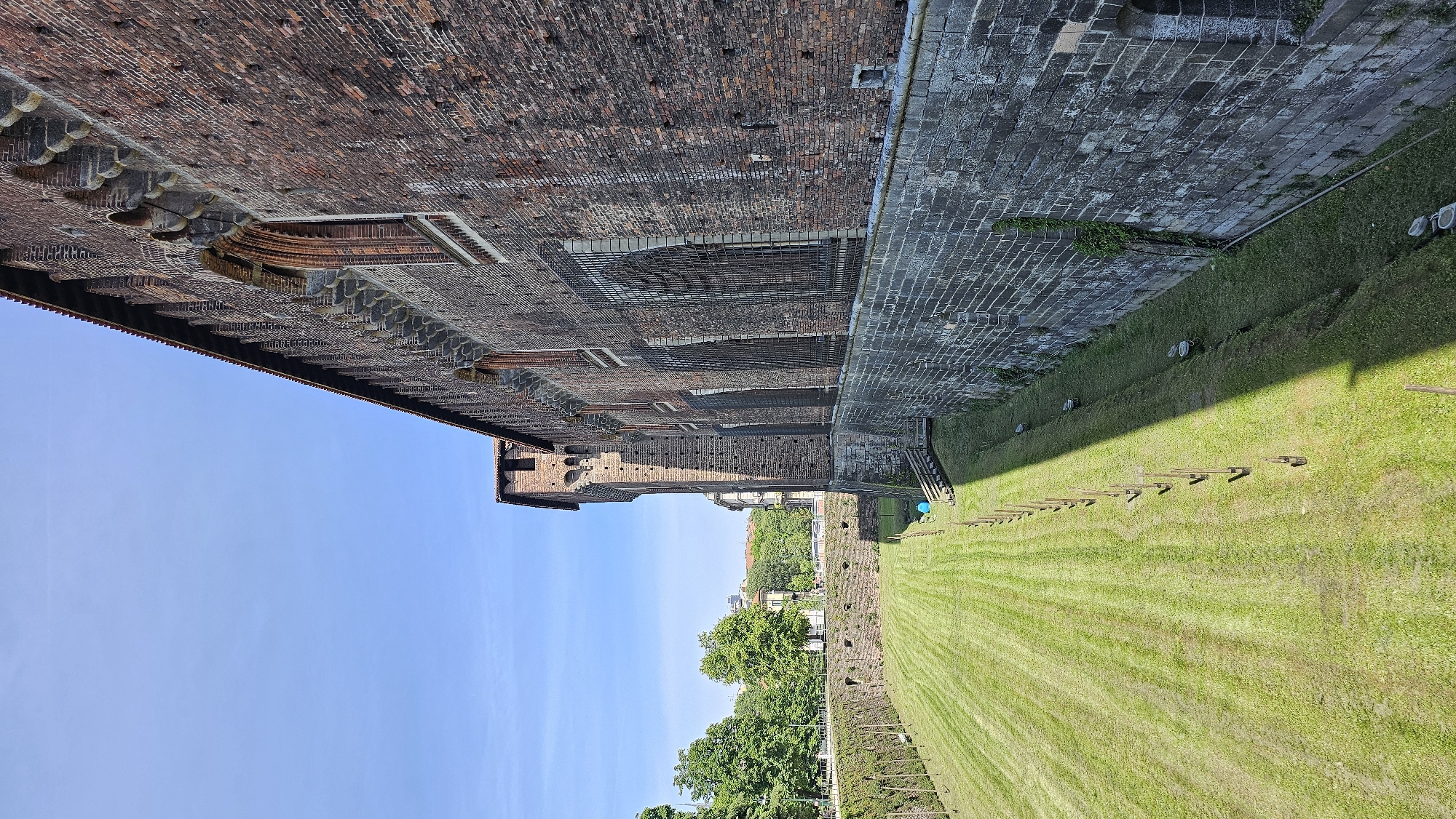 The image depicts the Sforza Castle in Milan, Italy, with a clear blue sky overhead. The castle's brick facade, featuring windows and decorative elements, extends across the upper portion of the frame. A grassy area is adjacent to the castle's wall, with wooden structures lining the edge of the grass. Further back, trees and buildings are visible in the distance, suggesting an urban setting. No people are visible in the frame.