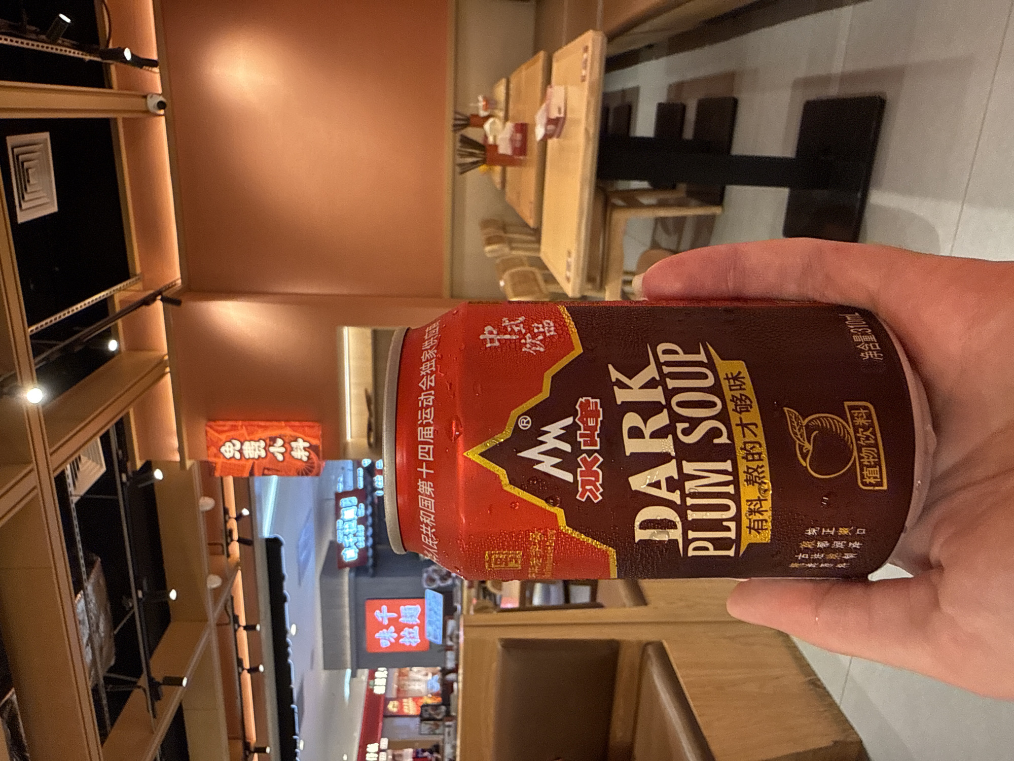 A person's hand holds a condensation-covered can of "DARK PLUM SOUP" inside what appears to be a restaurant. The can has a red and dark brown label with Chinese characters and a mountain logo. In the background, there are tables with chairs, lighting fixtures on the ceiling, and orange-colored walls. Some signage is visible in the distance, including a red banner with Chinese characters and a sign that seems to be for a noodle shop. The environment suggests an indoor dining establishment in a city.