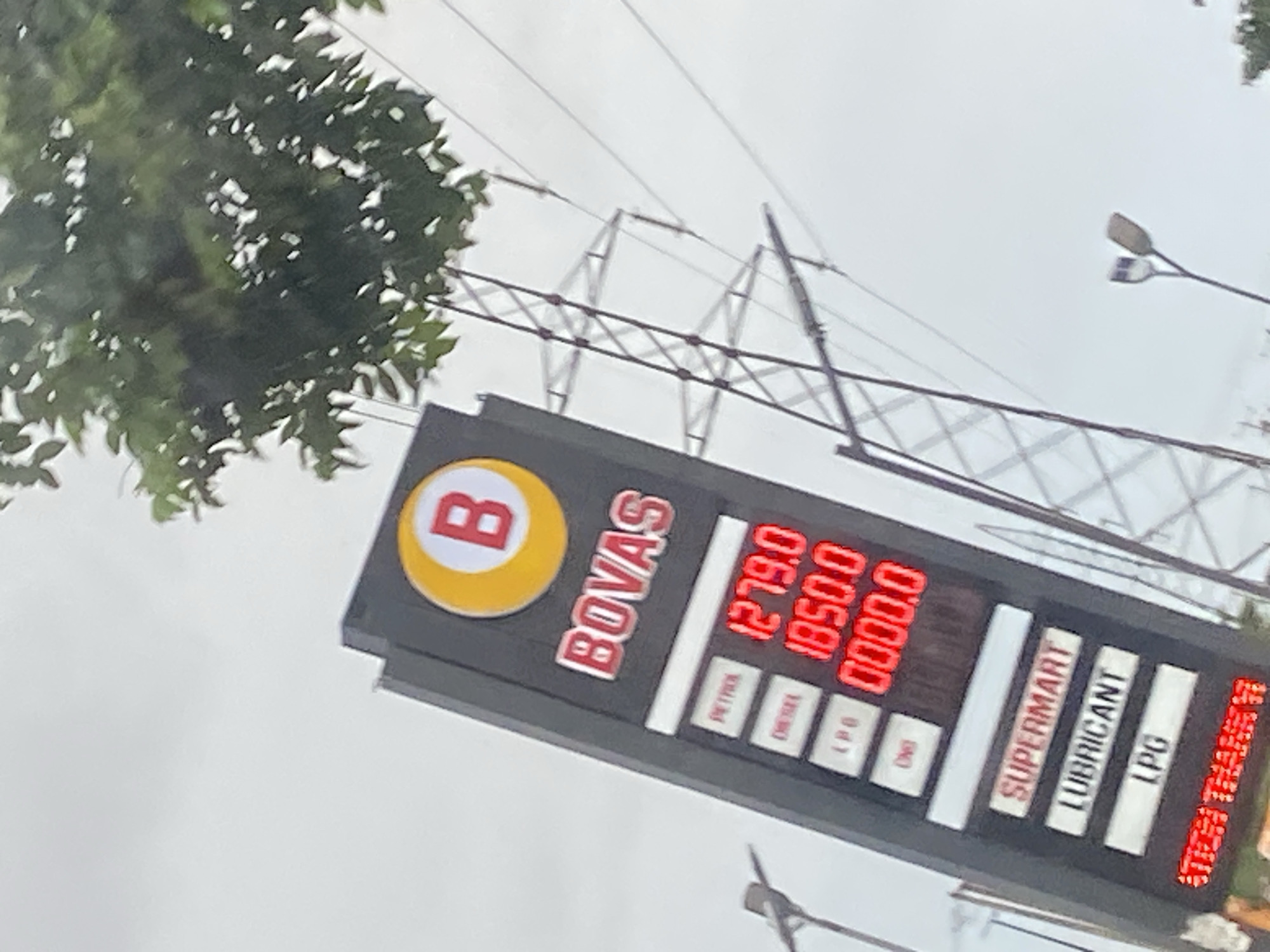 This image captures a close-up view of a BOVAS gas station price board in Ikorodu, Nigeria, against a backdrop of a cloudy sky, lush green trees, and power lines supported by a transmission tower. The prominent black price board features the BOVAS logo at the top, a red "B" within a yellow circle. Below the logo, the word "BOVAS" is displayed in red lettering with a white outline.
The price board lists fuel prices for different types of fuel. The first entry, labeled "PETROL," shows a price of "1279.0." Below this, "DIESEL" is listed with a price of "1050.0." Further down, "LPG" is shown with a price of "0000.0," and "CNG" is also present but its price is not clearly visible. Additional signage indicates "SUPERMART," "LUBRICANT," and "LPG" at the bottom of the board.
In the lower right corner of the image, a red LED display shows the text "OPEN THANKSGIVING."
The overall scene is typical of a street view in an urban or semi-urban area, with the gas station signage being the primary focus, juxtaposed against the infrastructure of power transmission and natural elements like trees. The image appears to be taken from a slightly low angle, looking up towards the price board and the sky.