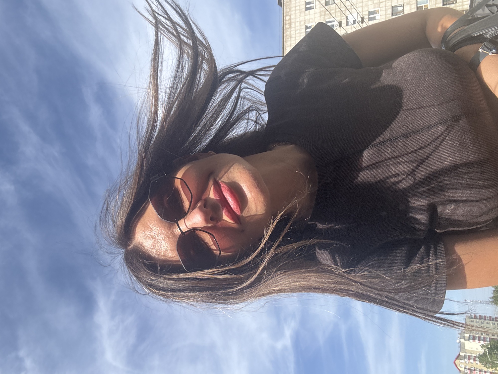 A young woman wearing sunglasses and a black t-shirt is positioned outdoors, facing the camera with a backdrop of a partly cloudy blue sky. Her long brown hair is windswept. The environment includes buildings suggestive of an urban landscape. A location in the city of Chaikovsky, Russia, is specified but not discernible from the image alone.