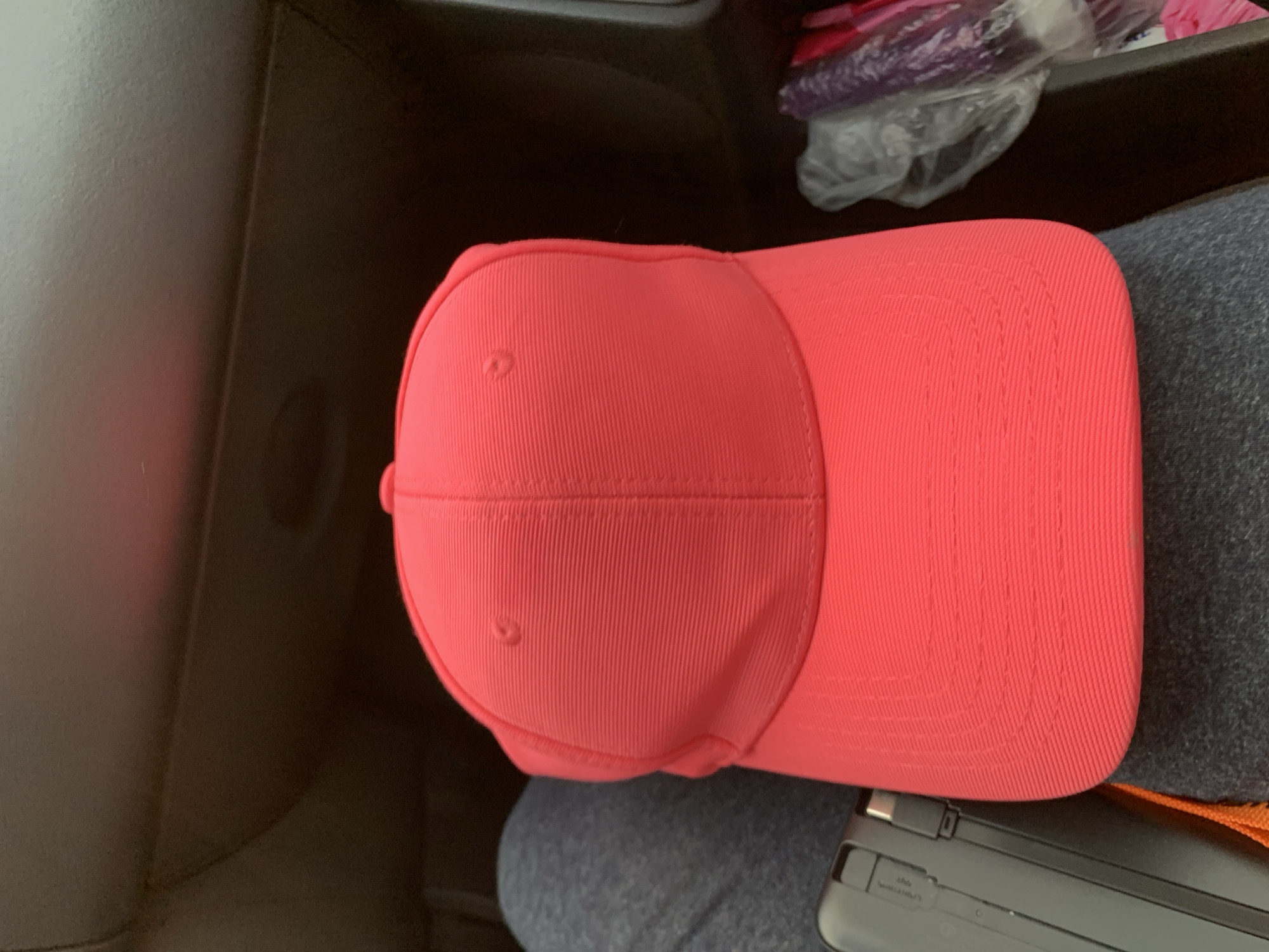 A close-up, overhead shot captures a bright coral pink baseball cap resting on a person's lap. The cap features subtle horizontal ribbing and a small button at the top. To the left of the cap, a black electronic device with a silver connector is visible. The background consists of dark, textured material, possibly the interior of a vehicle. To the right, a plastic bag contains purple and pink items, suggestive of toiletries or personal care products. The scene appears to be set in the rural area of Блеўчыцкі сельскі Савет, Belarus.