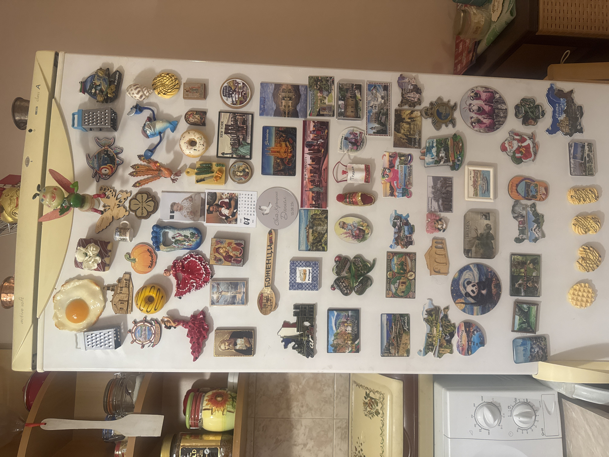 The image shows a white refrigerator covered in magnets. The magnets are of various shapes and sizes, and depict a wide range of subjects, including food, animals, landmarks, and cultural items. There are also some magnets with text, likely from different locations visited. The refrigerator is located in a kitchen, as evidenced by the presence of a microwave and kitchen cabinets in the background. The lighting suggests it is daytime, with artificial light also being used.