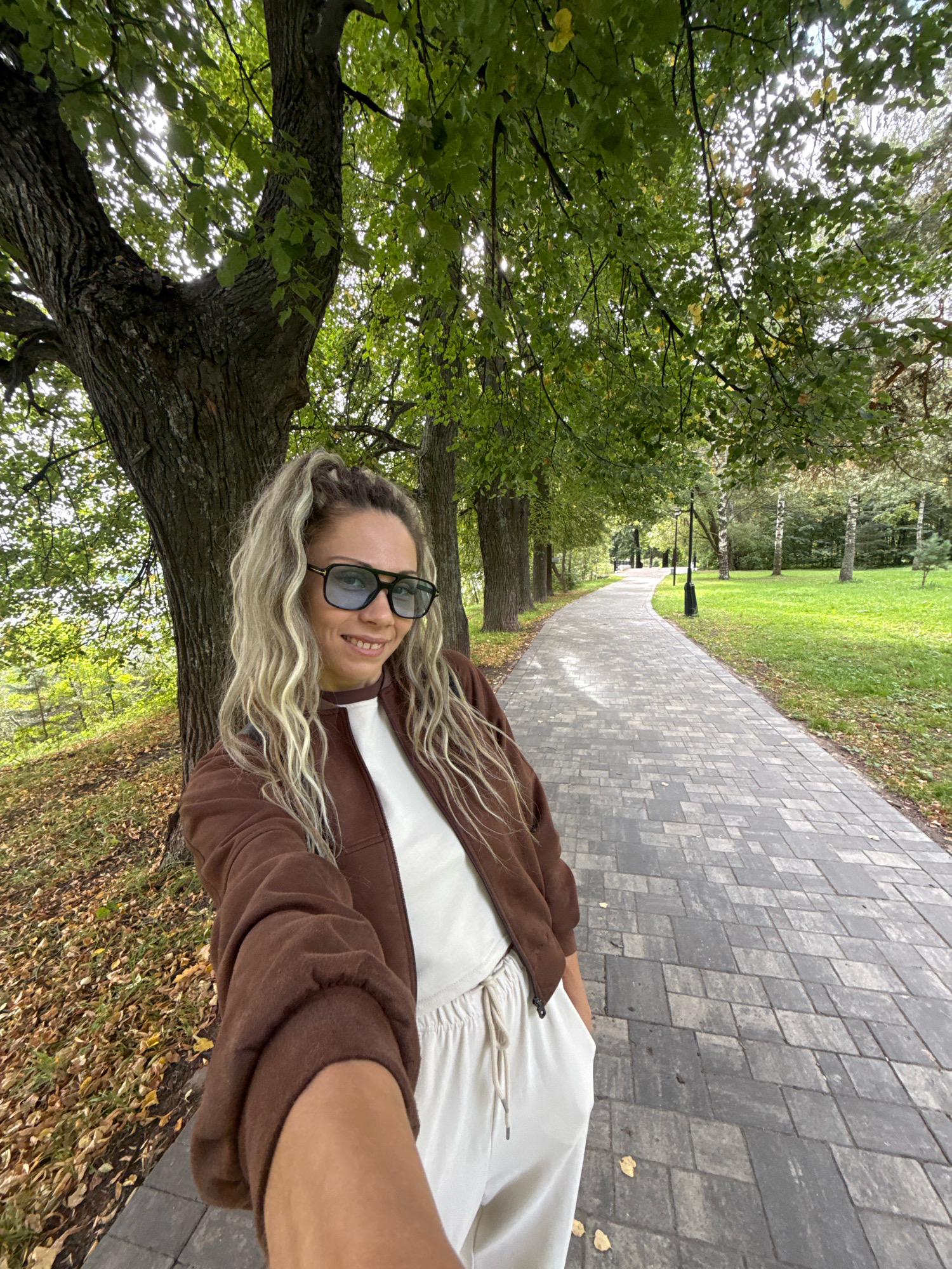 In Rusanovo, Russia, a blonde woman with curly hair tied in a ponytail stands in a park next to a tree, holding a camera to take a selfie. She wears a brown zip-up hoodie, a white top, and light-colored pants. The woman is smiling, and her sunglasses have a bluish tint. Behind her, a paved walkway stretches into the distance, flanked by trees and a grassy area. The ground is covered with fallen leaves, indicating it is autumn. The scene suggests a casual outdoor setting within the city of Rusanovo.