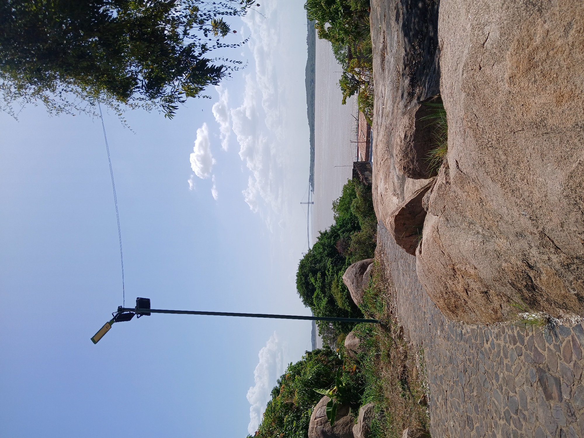 The image shows a scene on a cobblestone path bordered by large rocks and vegetation. A light pole stands to the left of the path, extending into a blue sky with scattered clouds. In the distance, a body of water can be seen with a bridge spanning across it. The location is in the city of Unknown, Unknown.