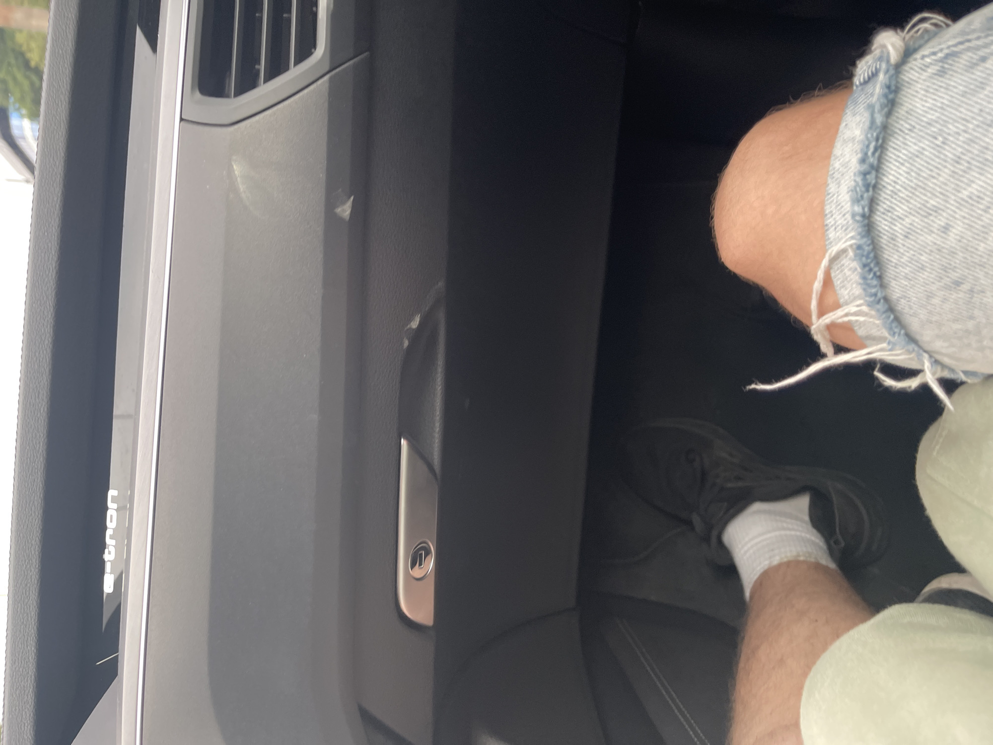 An interior view of a car's dashboard and a person's legs is depicted. The dashboard is black, with an "e-tron" logo visible on the upper section. A silver trim surrounds a recessed area, possibly a glove compartment, with a small latch or keyhole mechanism. To the right of this, a car vent with horizontal slats is present. Dust or smudges are visible on the dashboard surface. In the foreground, a person's legs are visible. The left leg is clad in a light green garment, and the right leg is covered by distressed denim jeans, showing rips and frayed edges around the knee. The person is wearing white socks and dark athletic shoes. The scene is shot from a low angle, looking upwards towards the dashboard. The background is dark, suggesting the car's interior. The image does not provide any information about the specific location within the city of Kyiv, Ukraine.