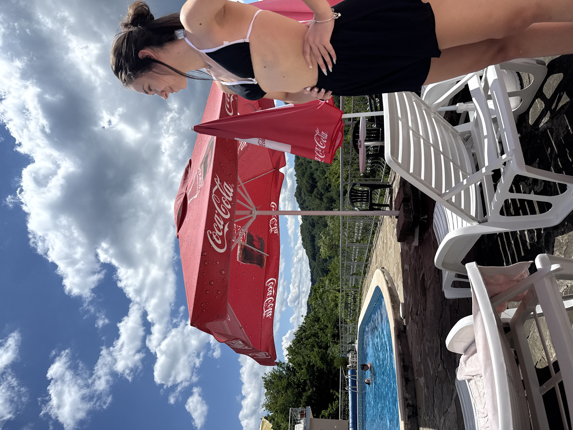 In Shipkovo, Bulgaria, on a sunny day with scattered clouds, a young woman stands by a swimming pool. She is wearing a black bikini top and black shorts, with her hands on her hips. A large red Coca-Cola umbrella is open behind her, and a smaller, closed red umbrella is next to it. A swimming pool with blue water is visible to the left, with two people swimming in it. White lounge chairs are scattered around the pool area. In the background, lush green trees cover rolling hills, and a building with a balcony is on the left.