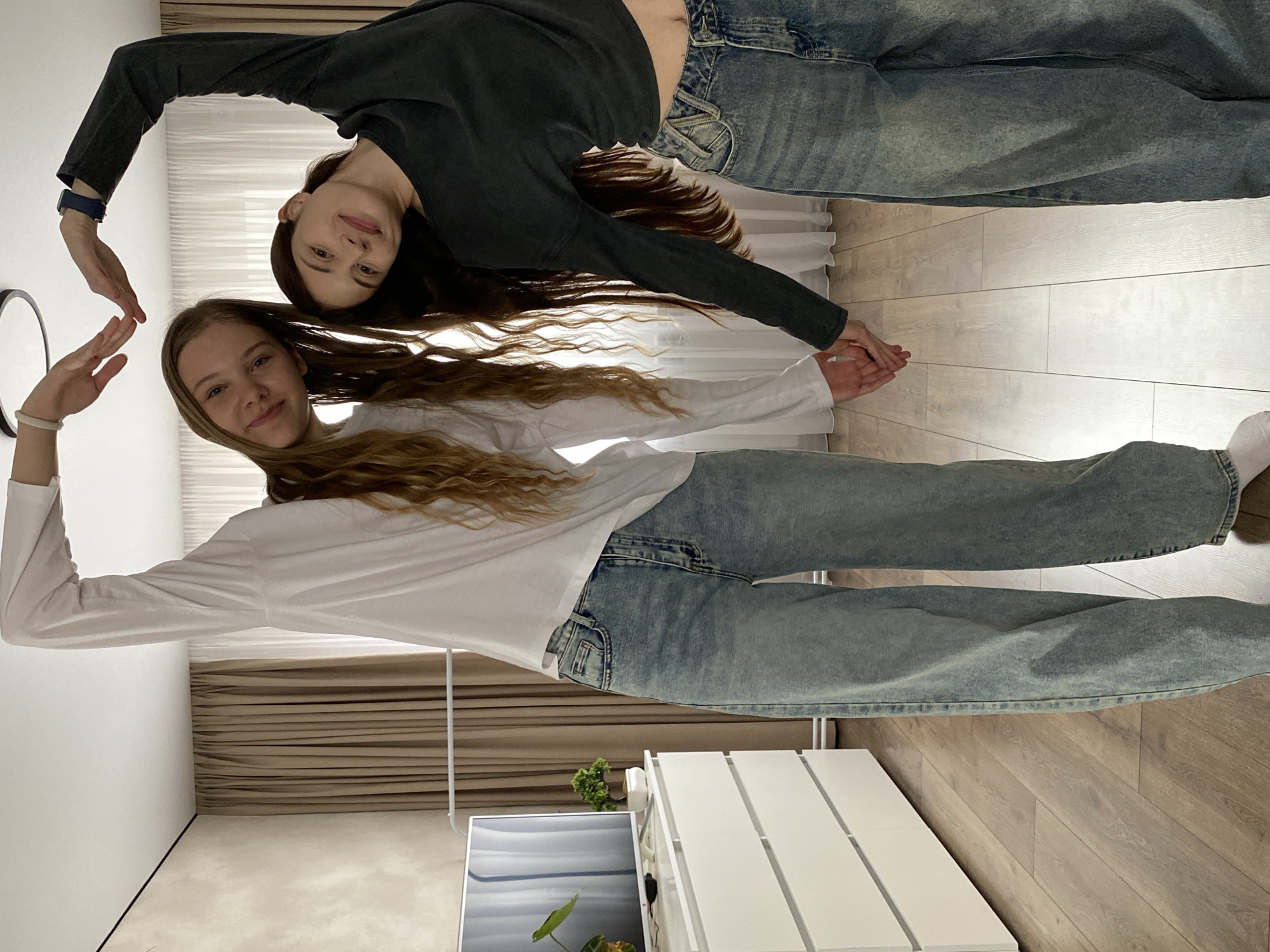 Two young women are posing on a wooden floor indoors. The woman on the top wears a long-sleeved black shirt and jeans, and the woman on the bottom wears a long-sleeved white shirt and jeans. They are holding hands in the middle. They are both forming the shape of a heart with their arms. A white cabinet, a plant, curtains, a mirror, and a monitor are in the background. The location is Votkinsk, Russia.