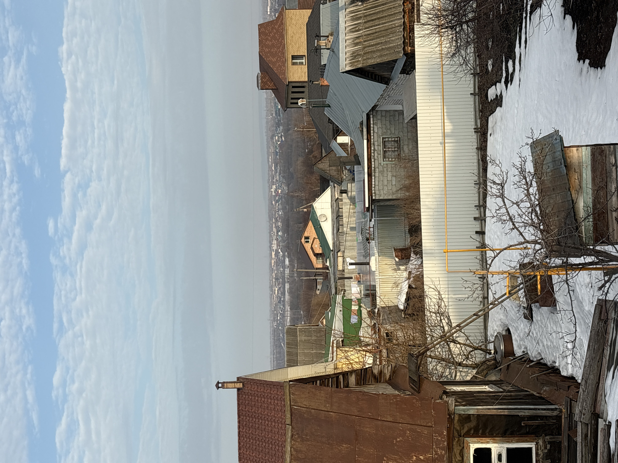 The image shows a view of a residential area in Ulyanovsk, Russia. It appears to be a mix of older, possibly decaying structures, and newer buildings. The foreground contains dilapidated, wooden houses with rusted roofs and window frames. In the middle ground, there are houses with various roof styles (metal, brick, and other materials). A white building with gas pipes is visible. Patches of snow cover the ground, indicating either a recent snowfall or the lingering effects of winter. In the background, a hazy urban skyline can be seen. The sky is partially cloudy. Overall, the scene suggests a post-Soviet urban landscape undergoing a transition.