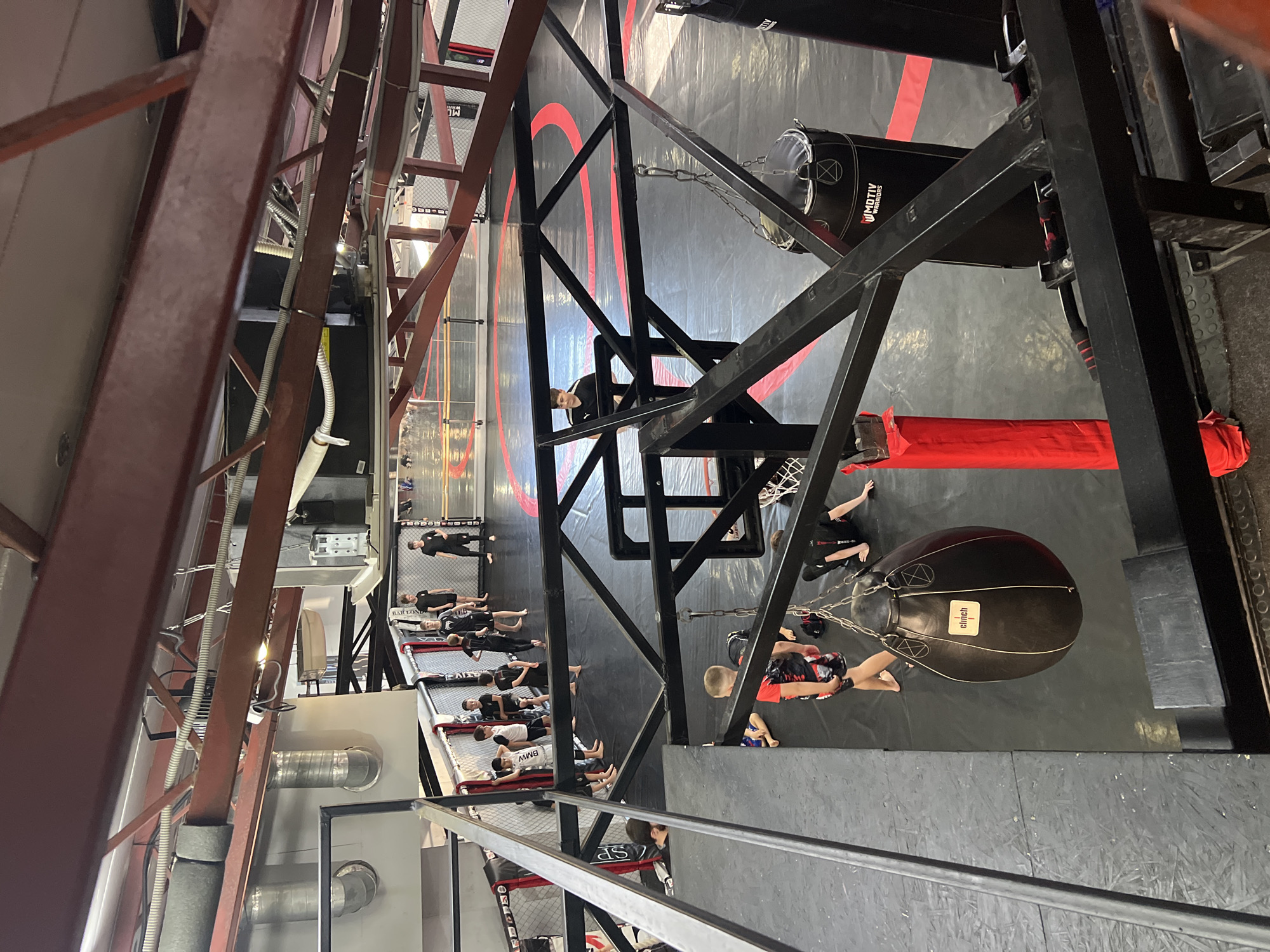 This photograph captures an indoor combat sports training facility, viewed from a high angle looking down into the space. The scene is dominated by black floor mats with prominent red markings, and robust metal support structures that crisscross the frame.
Numerous individuals, mostly young people or children, are present in various areas of the gym. To the left side of the image, a group is visible inside a black-caged ring or training area, lying on their backs on the mats, possibly resting or performing floor exercises. One person's shorts clearly show the text "BMW".
In the upper-middle section of the image, a person in black clothing stands within a large red circular design on the floor. To the right, several punching bags hang from black metal frames. A large, black, teardrop-shaped punching bag with "clinch" written on it is prominent in the foreground, near a person wearing red and black shorts. Further back, another black cylindrical punching bag is visible, featuring the text "MOTIV WARRIORS".
The gym's infrastructure includes thick red and black metal beams and trusses that form part of the building's support system and equipment mounts. Red padded barriers are placed along the floor beneath some of these structures. Exposed ventilation ducts and electrical conduits are also visible running along the metal framework.
The overall setting is that of an active training environment in a combat sports gym located in Sochi, Russia.