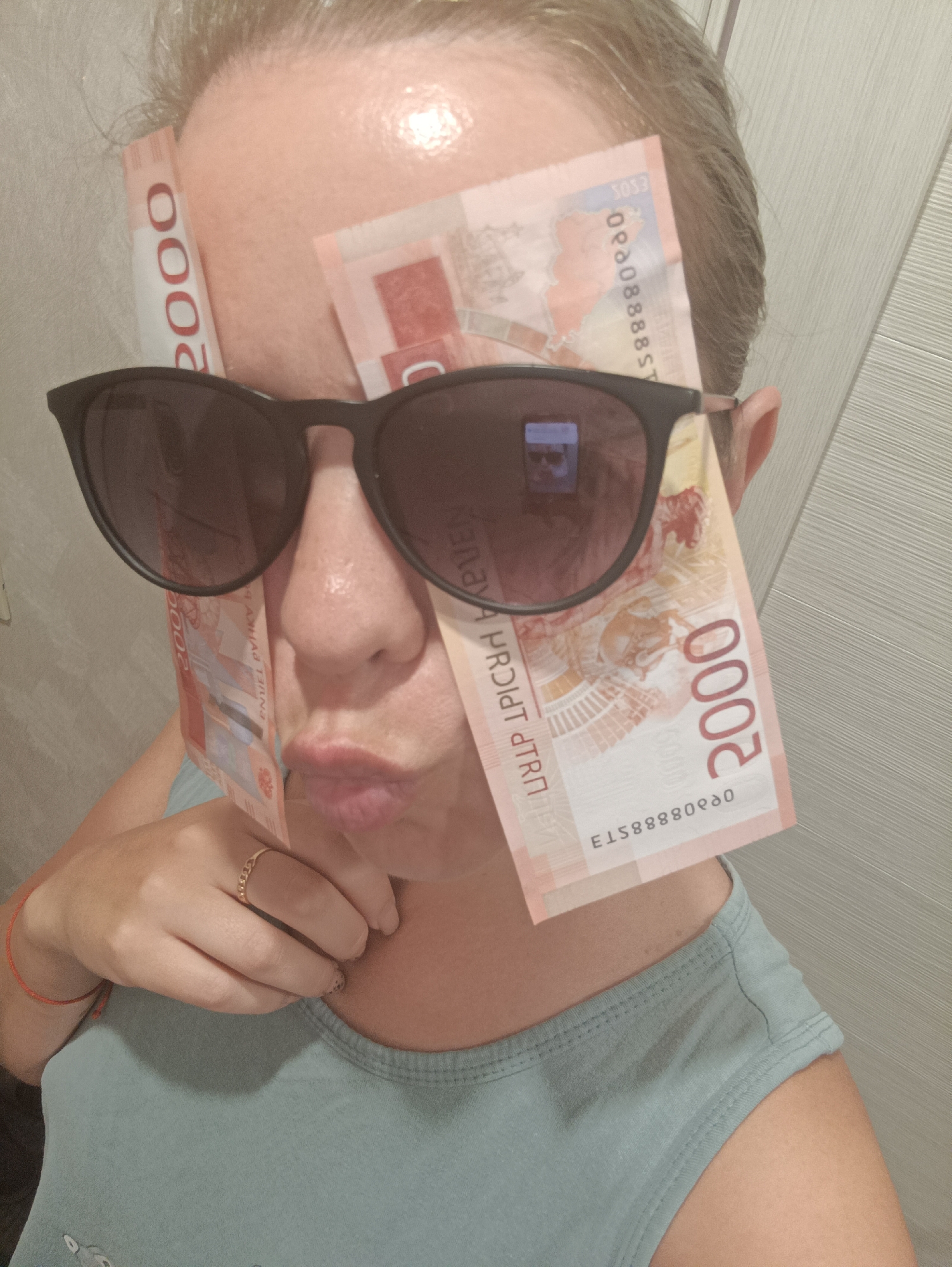 A person with fair skin, graying hair, and wearing black sunglasses poses with several Russian 2000-ruble banknotes strategically placed on their face. The bills partially obscure their face, one positioned vertically on the left, another at an angle across the forehead, and one draped down the right side of their face. The person's lips are puckered. The person wears a light blue t-shirt and a ring on their left hand. Based on the banknotes present in the image, this photograph was taken in Russia, but it is NOT possible to tell if this photograph was taken in the city of Chaikovsky, Russia. The background is a plain, light-colored wall with a vertical linear texture.