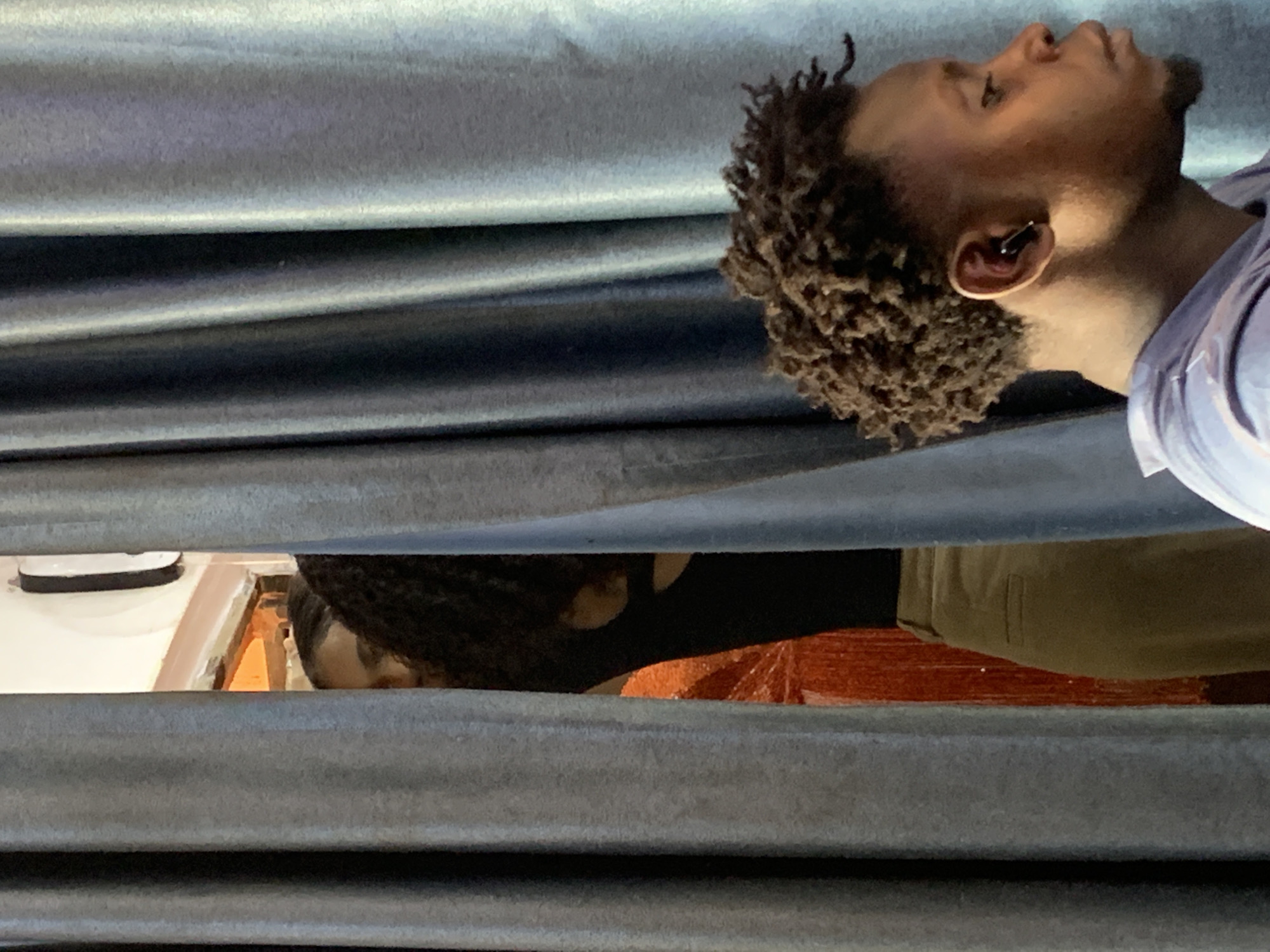 The image shows two individuals. One, located in the upper right quadrant, is a Black male with short, curly hair and an earbud in his right ear. He is wearing a light blue shirt. His head is tilted back.
The second individual is located in the center of the image, visible through a gap in what appears to be corrugated metal siding. Only the back of their head and shoulder are visible. They have dark hair and are wearing a black shirt and olive-colored pants. Their arm is extended, perhaps resting on a red-brown surface.
The environment is characterized by corrugated metal siding, with the second individual visible through a horizontal gap in the siding. The exact location within Sabojibi, Nigeria is not discernible from the image. No specific actions or interactions are evident.