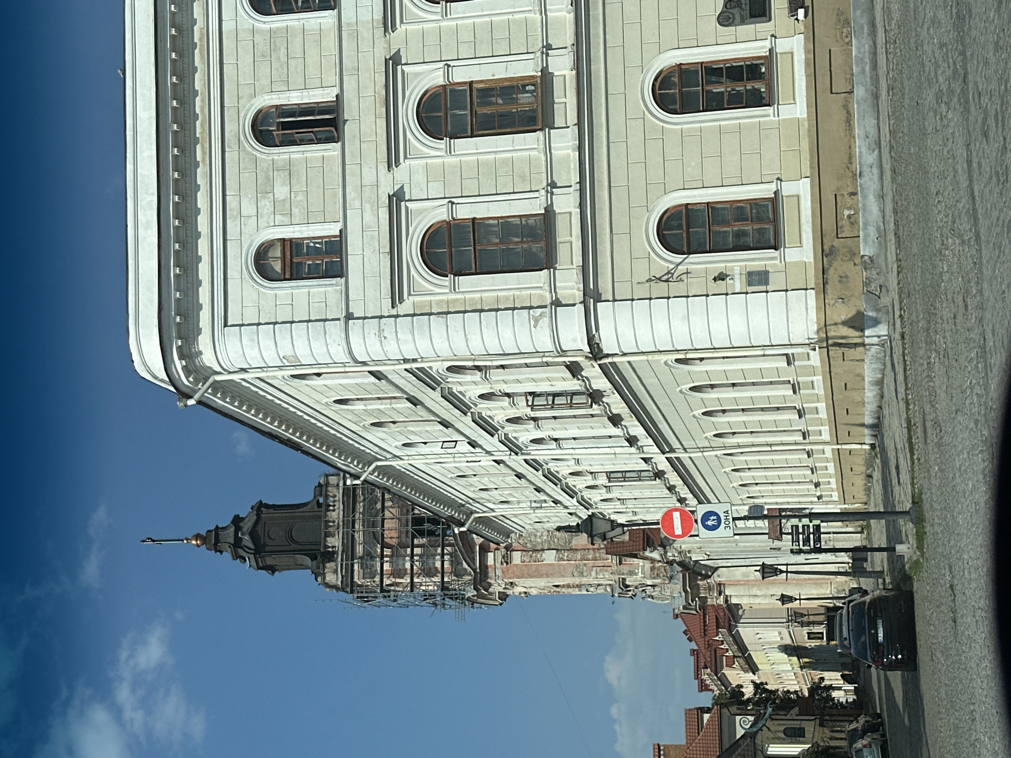 The image captures a street scene in Kamianets-Podilskyi, Ukraine, focusing on the architecture and urban environment. In the foreground, a cobblestone street is visible, with a black sedan parked on the left side. To the right, a light-colored, multi-story building with arched windows dominates the frame, its facade adorned with decorative moldings. Further in the background, a tall, dark tower with scaffolding and construction elements rises against a bright blue sky with scattered clouds. Other buildings with red-tiled roofs line the street to the left, creating a historic town atmosphere. Traffic signs, including a red "no entry" sign and a blue sign with pedestrian figures and "ЗОНА" (Zone), are present on a pole. The lighting suggests it is daytime, likely with sunlight casting shadows.