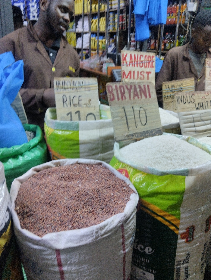 Bags of rice and beans for sale with hand-written signs. One sign reads "INDIAN RICE" and another says "BIRYANI" with a price of "110" (presumably currency). A sign says "KANGORE MUST". The grains are in large sacks, with two people behind the merchandise. Shelves of various items line the background, indicating a small store or market setting.