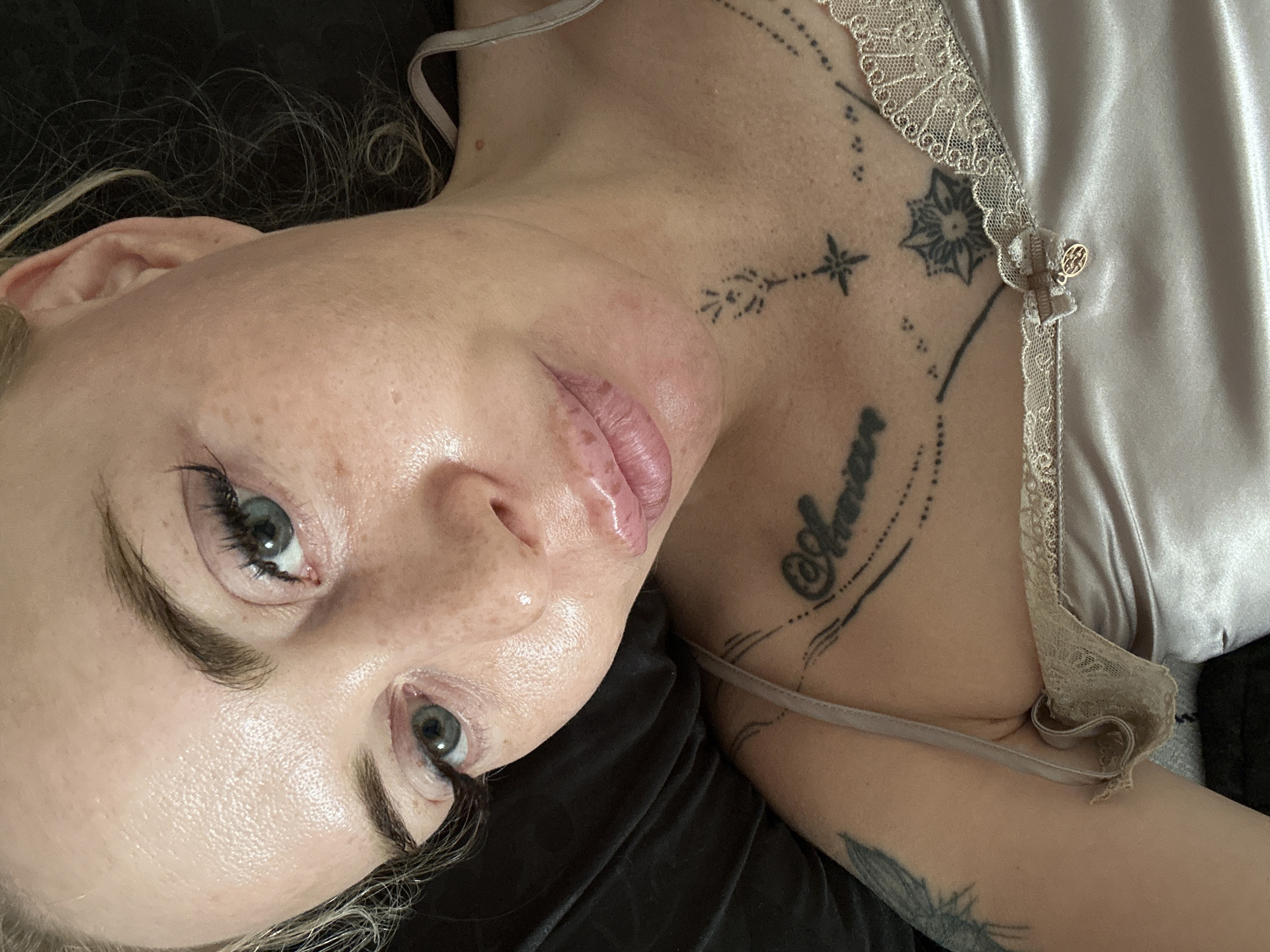 A close-up, overhead shot shows a fair-skinned person lying down and looking up towards the camera. The person has blue-green eyes, dark eyebrows, and light blonde hair. The person is wearing a light pink silk camisole with lace trim. There are several tattoos visible on the person's chest and shoulder, including script, a star, and what appears to be a butterfly. The background is dark, suggesting a dimly lit indoor environment, possibly a bedroom. The image is taken from a high angle, with the person's face dominating the frame. The lighting is soft, highlighting the person's skin and makeup.