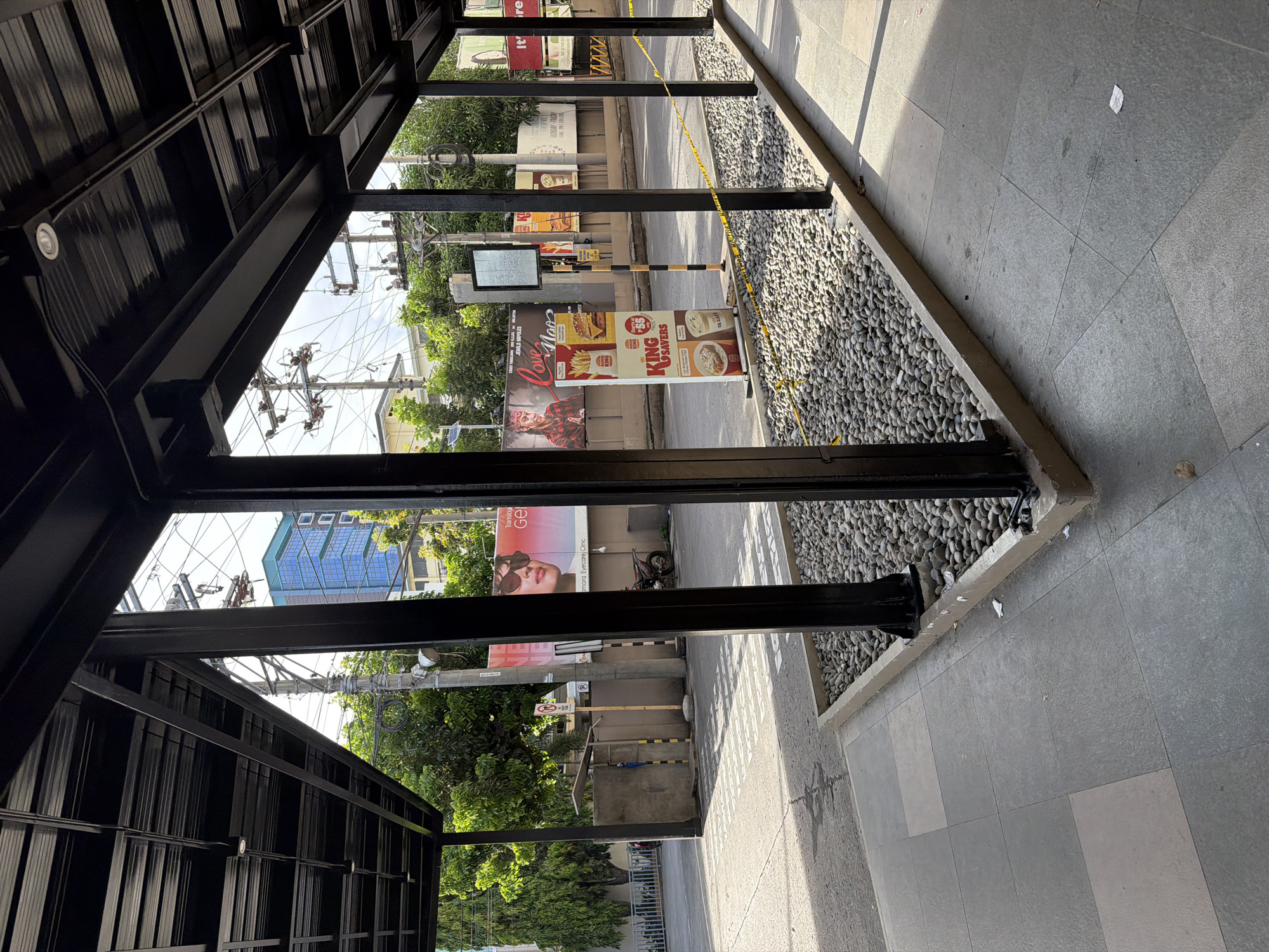 This outdoor, medium shot shows a covered walkway in Davao City, Philippines. The walkway, constructed from dark steel, provides a partial view of the street.
Advertisements and signage are visible across the street. These include ads for "Transitorio GE," "King Savers," and "Amora Eyecare Clinic." A pedestrian barricade with yellow and black stripes stands on the sidewalk, with yellow caution tape strung across.
The sidewalk is tiled in gray, with a rock-filled trough lining the edge. Power lines and greenery fill the spaces above the buildings across the street. The photo appears to be taken during the day, with ample sunlight.
