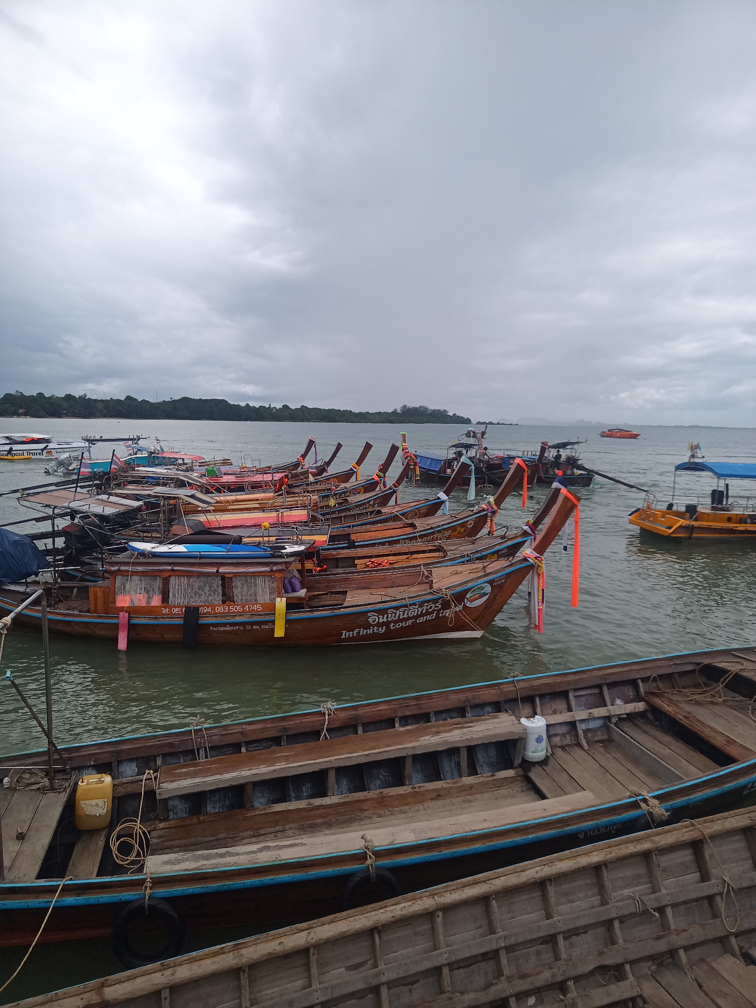 This image shows a collection of longtail boats docked in Ao Nam Mao, Thailand, under a cloudy sky. The boats are primarily brown with colorful accents, and some have signage in Thai and English. One boat reads "Infinity Tour and tre" and includes a phone number. Some boats have orange ribbons hanging from their prows. In the background, an island is visible across the water. The foreground shows a partially visible wooden boat with blue accents.