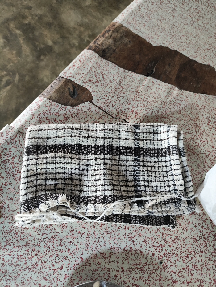 A folded, black and white plaid fabric, likely a towel or dishcloth, is the primary subject, resting on a patterned surface. The fabric has a classic checkered design, with visible fraying and loose threads along one of its folded edges.
The patterned surface underneath is a light-colored material, possibly a tablecloth or mat. It features a speckled texture of small reddish-brown dots scattered across its lighter background. Interspersed within this speckle pattern are larger, irregular brown shapes that resemble abstract natural forms such as decaying leaves or organic smudges.
In the upper background and top left corner, a darker, plain, and slightly textured surface is visible, contrasting with the lighter tones of the foreground. The image is a static shot of household items. No human subjects are present, and no specific activity or event is taking place. The scene appears to be indoors. There are no discernible cues for time of day or weather, nor are any emotions conveyed. No visible text is present in the image. The general setting is consistent with a domestic environment in Selagai Lingga, Indonesia.