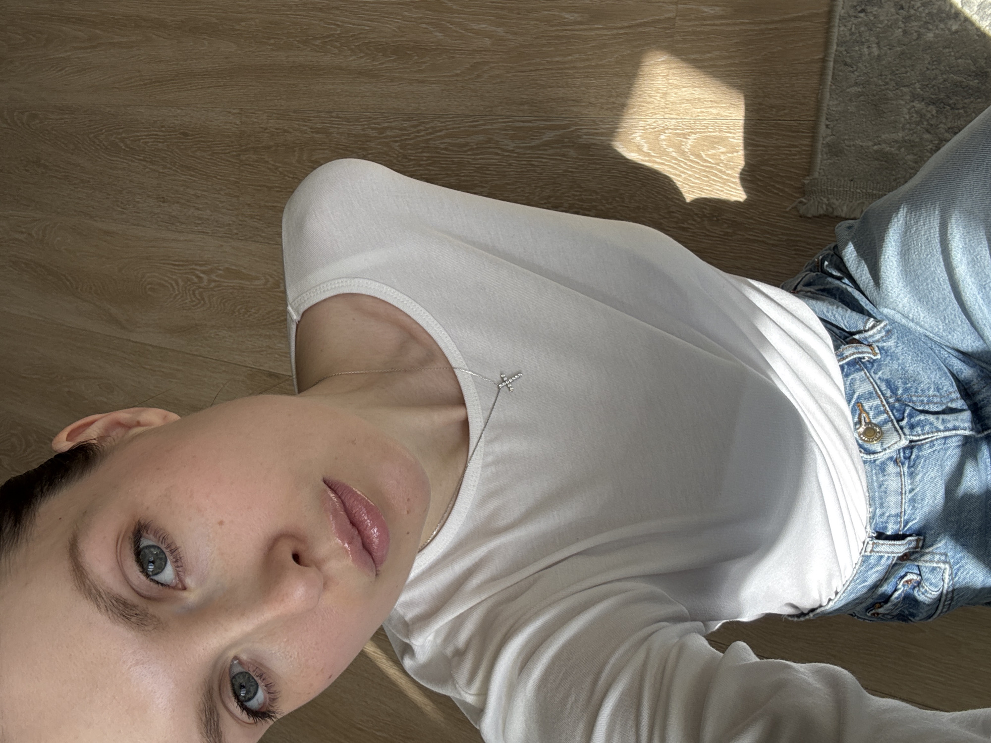 A young, fair-skinned person with gray eyes and dark hair is lying on a wooden floor. They are wearing a white long-sleeved shirt and blue denim jeans. A delicate silver necklace with a cross pendant is visible around their neck. Sunlight is illuminating the floor and casting shadows in the upper right portion of the image. A gray rug is partially visible in the upper right corner. The location and city are not indicated in the image and therefore cannot be determined.