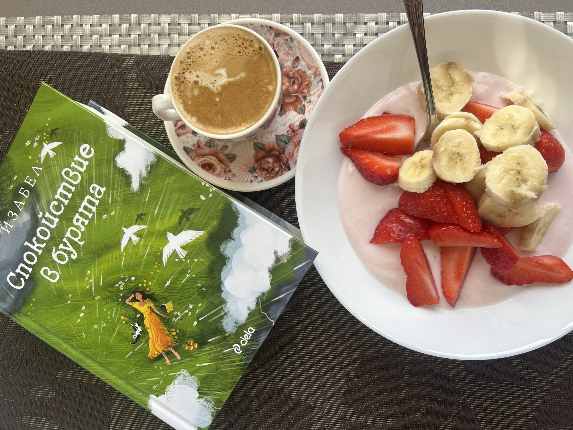 The image depicts a tabletop setting with a book, a cup of coffee, and a bowl of fruit. The book, titled "Спокойствие в бурята" (translated to "Peace in the Storm"), has a green cover with an illustration of a woman lying in a field. Next to the book is a cup of coffee with a floral saucer. A white bowl filled with yogurt, sliced strawberries, and sliced bananas sits to the right. A spoon rests in the bowl. The surface beneath the items is a woven placemat with a brown and white pattern. The photo appears to be taken in Tryavna, Bulgaria, based on the Bulgarian text on the book.
