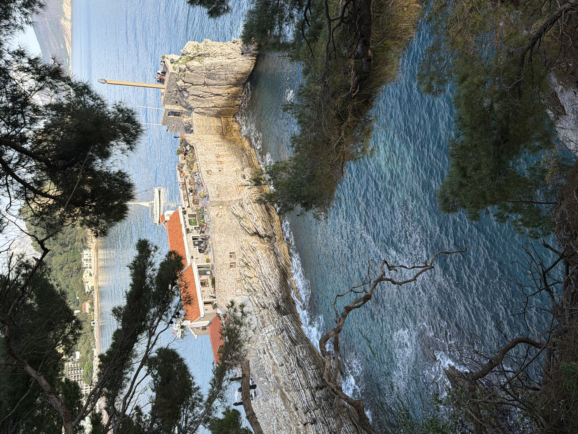 The image shows a coastal scene in Petrovac centar, Montenegro, from an elevated perspective. The blue sea is visible, with waves breaking against a rocky shoreline. A small island or peninsula is connected to the mainland by a causeway. A stone building with a red-tiled roof is situated on the peninsula. People are visible on a rocky outcrop at the end of the peninsula. Trees and vegetation frame the scene.