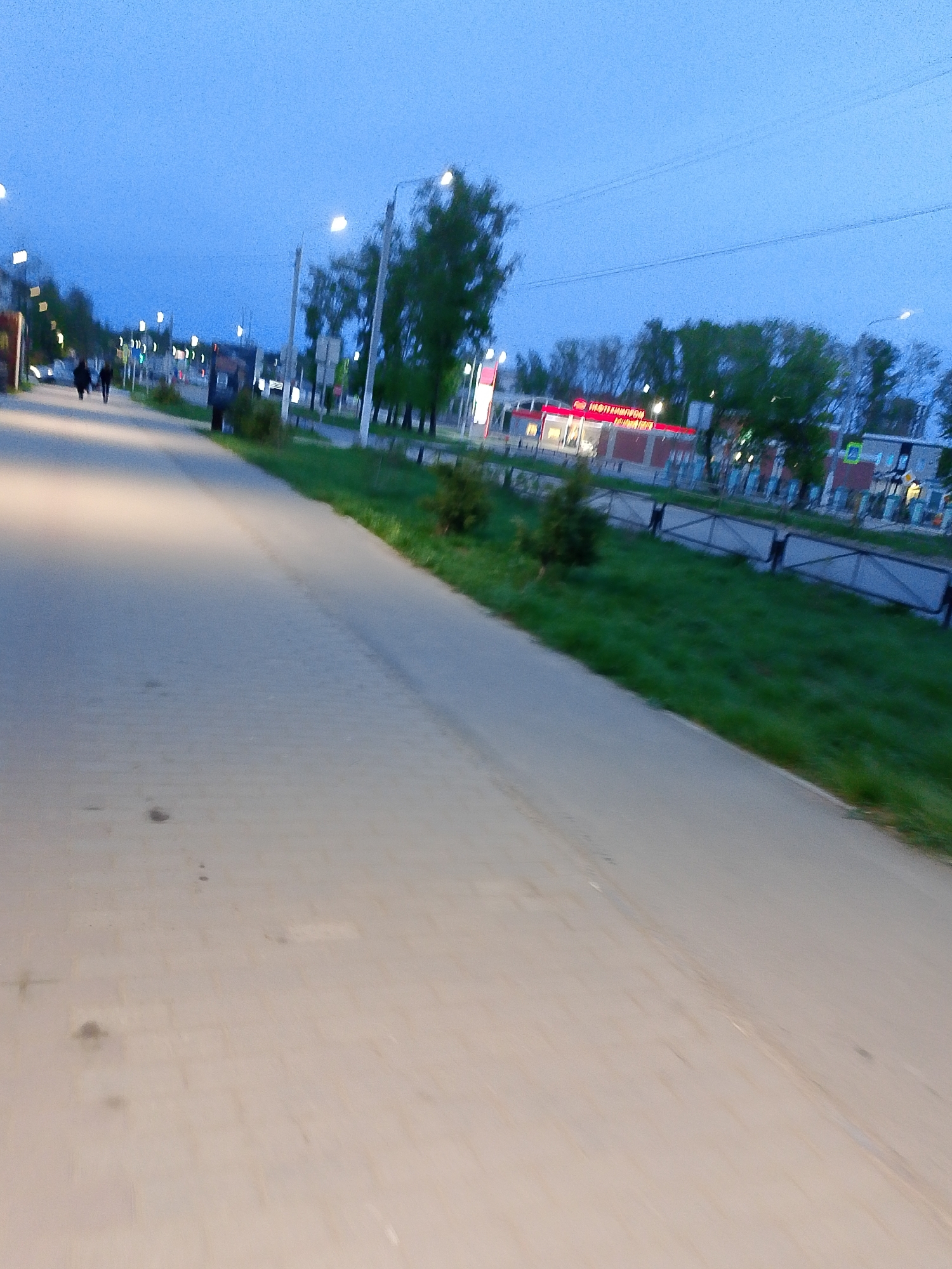 The image depicts a street scene in Chaikovsky, Russia, at dusk. A paved sidewalk stretches into the distance on the left, with a grassy strip separating it from the road. Trees and buildings line the right side of the road, including what appears to be a brightly lit commercial establishment in the distance. Two pedestrians are visible walking along the sidewalk in the distance. Streetlights illuminate the scene.
