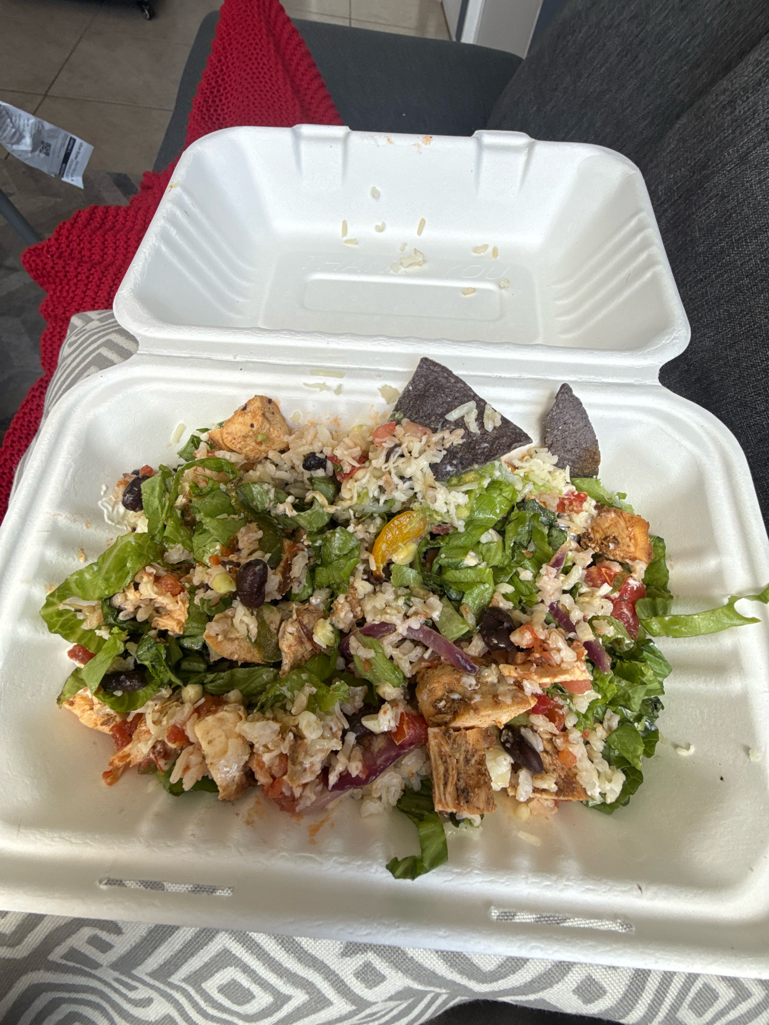 A white take-out container filled with a salad is shown from a top-down perspective. The salad contains chopped lettuce, rice, black beans, diced tomatoes, shredded red onion, chunks of grilled chicken, and two blue corn tortilla chips. The container is open, with the lid propped open and resting behind the main container. A red knitted blanket is visible behind the take-out container. The container is sitting on a gray and white patterned surface, and a dark gray couch is visible in the background. The scene appears to be indoors, possibly in a home. The location in Puebla, Mexico is not specified in the image.