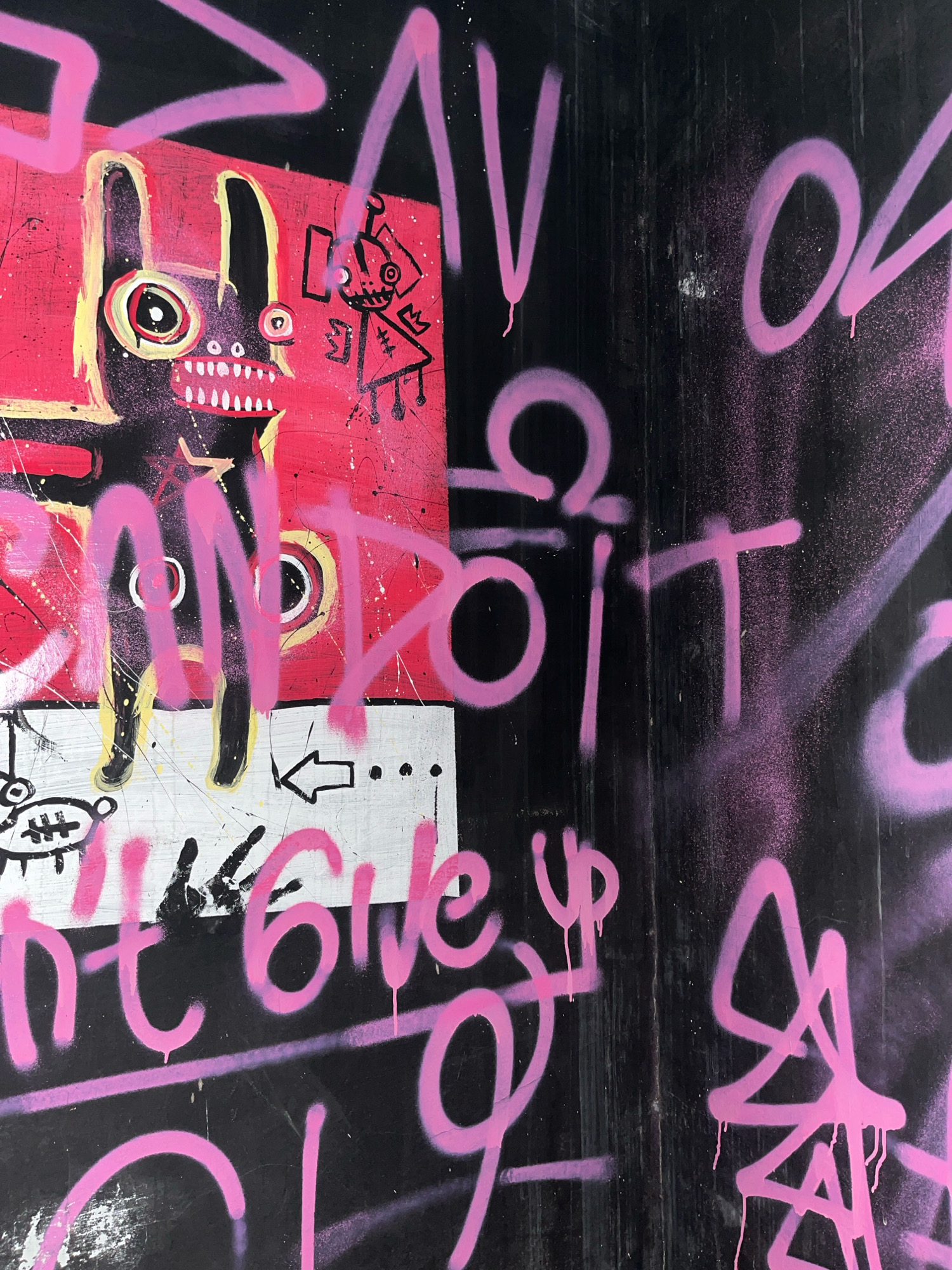 The image displays graffiti art on a black, textured surface, possibly a wall or a door, located in the city of Surakarta, Indonesia. The artwork features a cartoonish rabbit-like figure with wide, staring eyes and sharp teeth, rendered in black and yellow on a pink background. This figure is accompanied by a smaller, robot-like character with a crown. Pink spray paint is used extensively, forming bold, angular strokes and lettering throughout the composition. Some of the visible text appears to read "DON'T GIVE UP" and other abstract shapes. The overall style is energetic and somewhat chaotic, with drips of paint evident, suggesting a street art aesthetic. The black background exhibits vertical lines and some wear, indicating exposure to the elements.