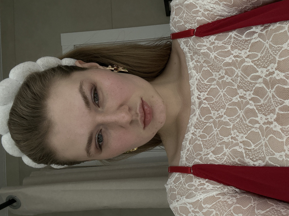 A woman with fair skin, blue eyes, and light brown hair looks directly at the camera. She wears a white, lacy top with red straps and a large, white, fluffy headband. Gold earrings are visible on both ears. The background is blurred, but a white doorframe is visible. The image is slightly rotated, giving it a tilted perspective.