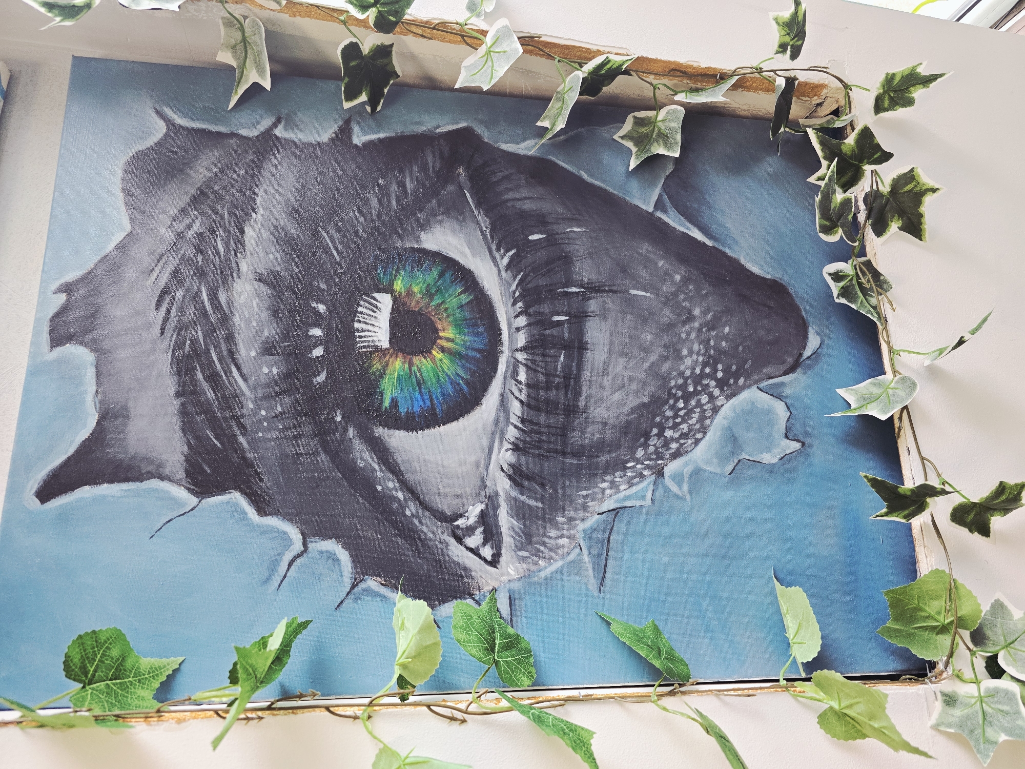 The image shows a painting of an eye tearing through a blue background. The eye is predominantly gray with a vibrant, multicolored iris (green, yellow, and blue). The painting is framed and decorated with artificial ivy. The location in Veliko Tarnovo, Bulgaria, cannot be determined from the provided image.