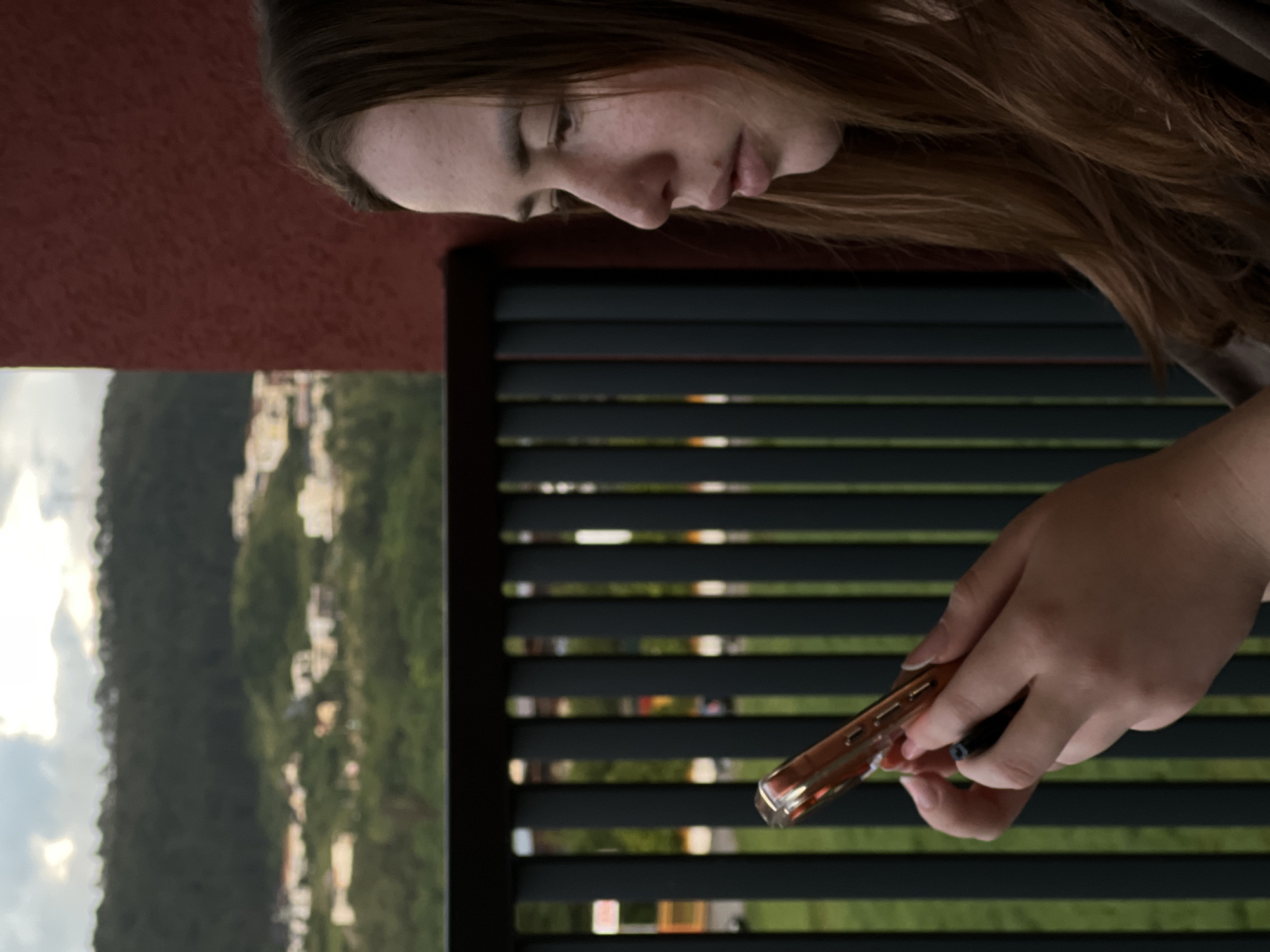An extreme close-up, shallow depth of field shot shows a young Caucasian woman with long, auburn hair from the chest up, looking down and to the left at a cell phone held in her hands. Her face is partially obscured by her hair. The woman is wearing a dark green t-shirt. The background consists of a dark green wooden fence, a red stucco wall on the right, and a blurred landscape with houses, trees, and a cloudy sky in the distance. This scene is likely captured on a balcony or patio overlooking a hillside residential area. The specific location in Florești, Romania, is not evident from the visual information provided in the image.