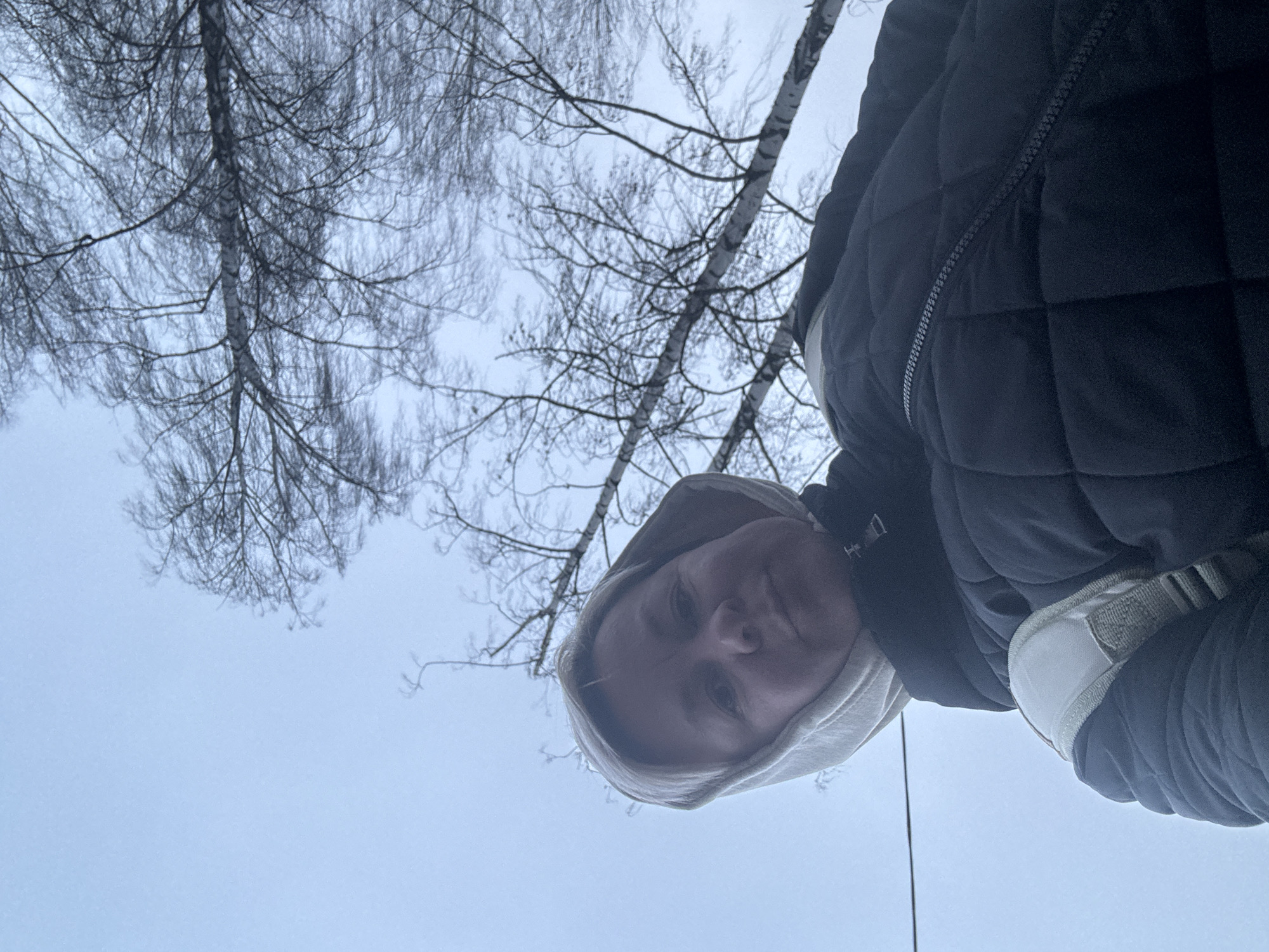 A low-angle shot captures a person wearing a hooded sweatshirt and a dark, quilted jacket. They are standing outdoors. Bare trees with white bark are visible against a pale sky. The location is Chaikovsky, Russia.
