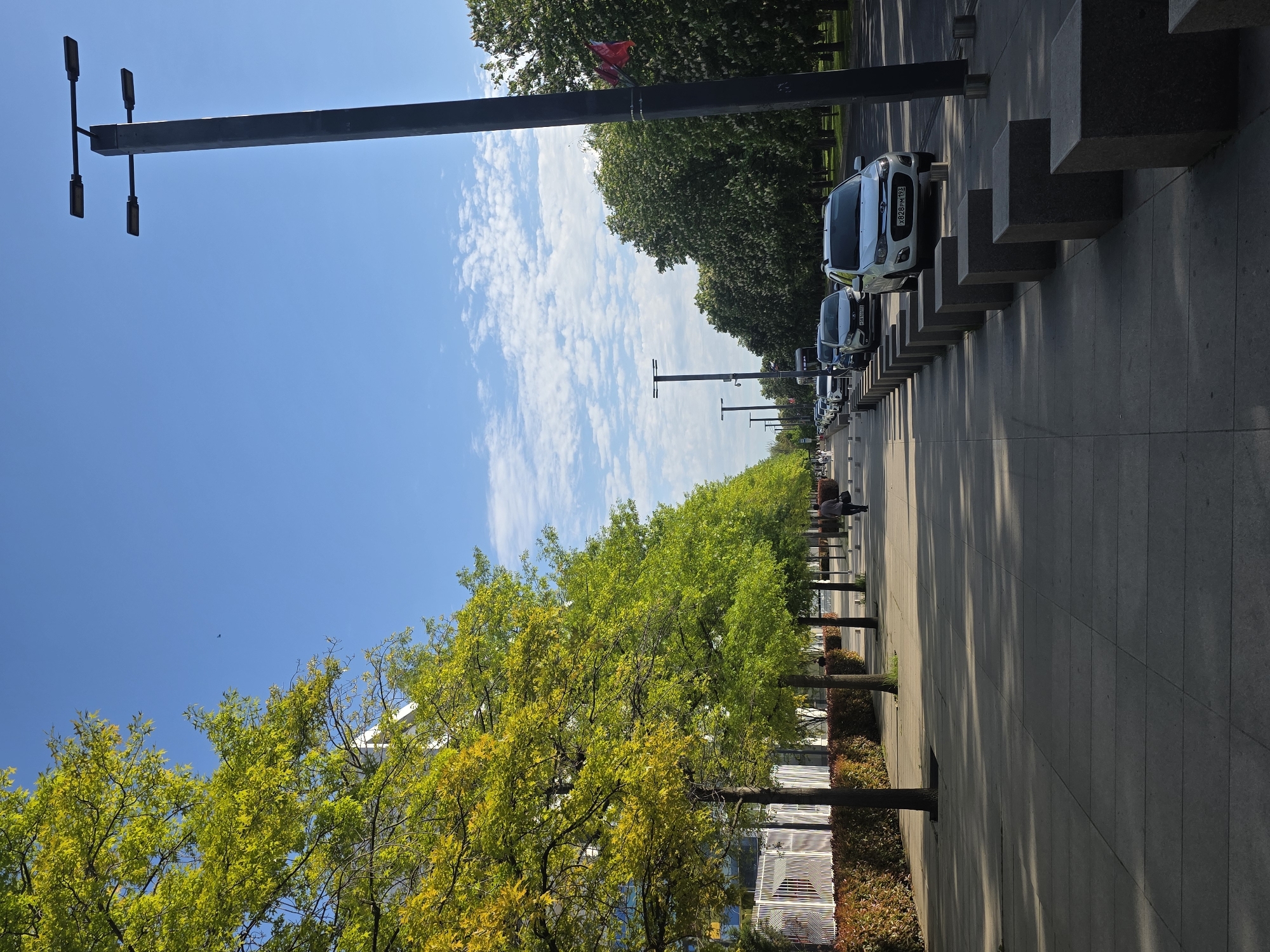 The image shows a street in Krasnodar, Russia, lined with trees on one side and parked vehicles on the other. The sky is blue with scattered clouds. There are street lights lining the road. Two people are seated on a bench. Adjacent to the row of parked vehicles are cubical gray concrete-like structures.