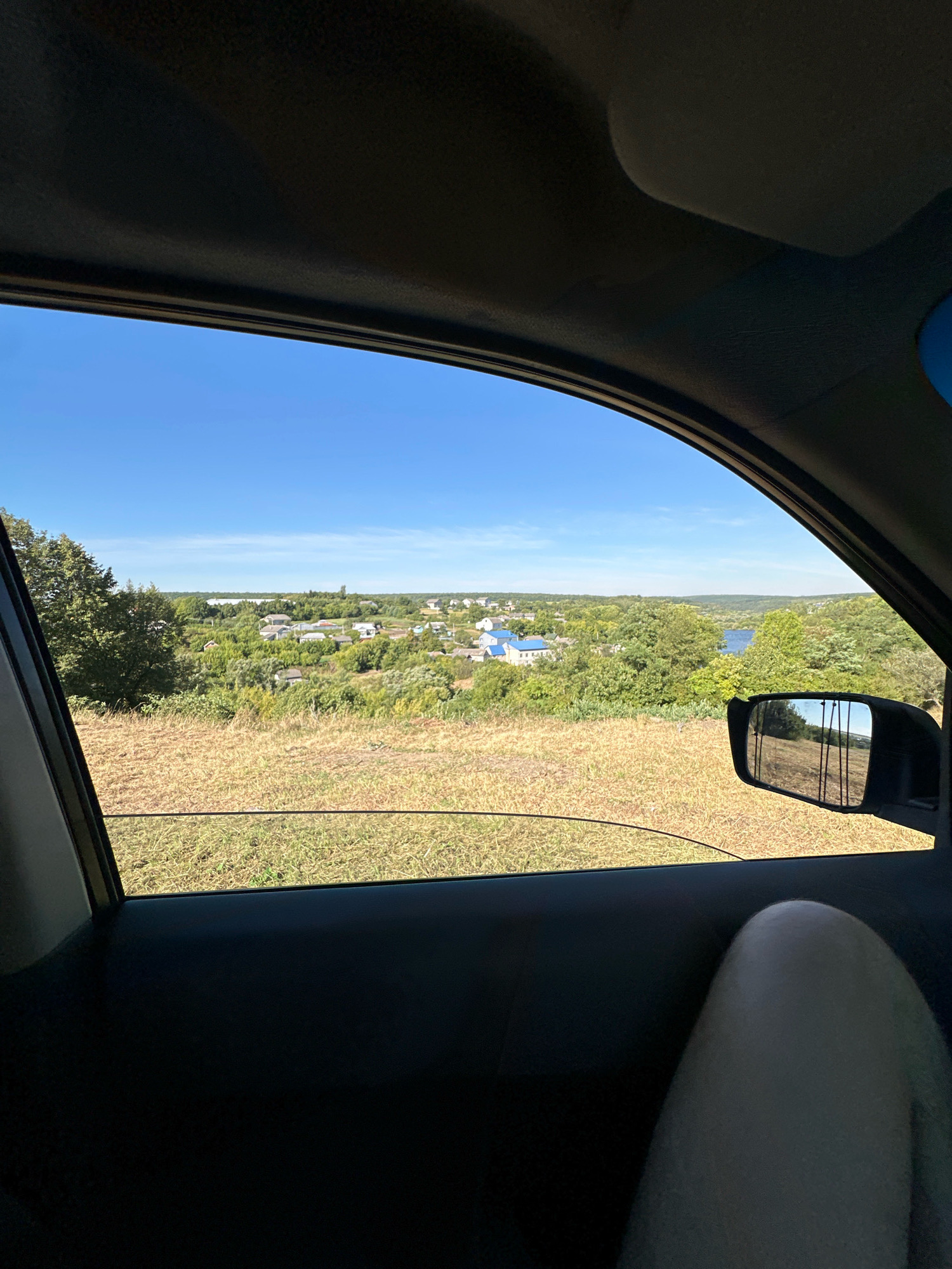 The image is taken from inside a vehicle, looking out through the passenger-side window. The view shows a rural landscape with a village nestled among green trees and rolling hills. Several houses with blue roofs are visible in the village. A body of water, possibly a river or lake, can be seen in the distance. The foreground consists of dry, brown grass and a car door interior. A person's leg, illuminated by sunlight, is visible in the lower right corner of the frame. A side mirror reflects a portion of the scene, including a fence or posts. The sky is clear and blue. The location is identified as Makiv, Ukraine.