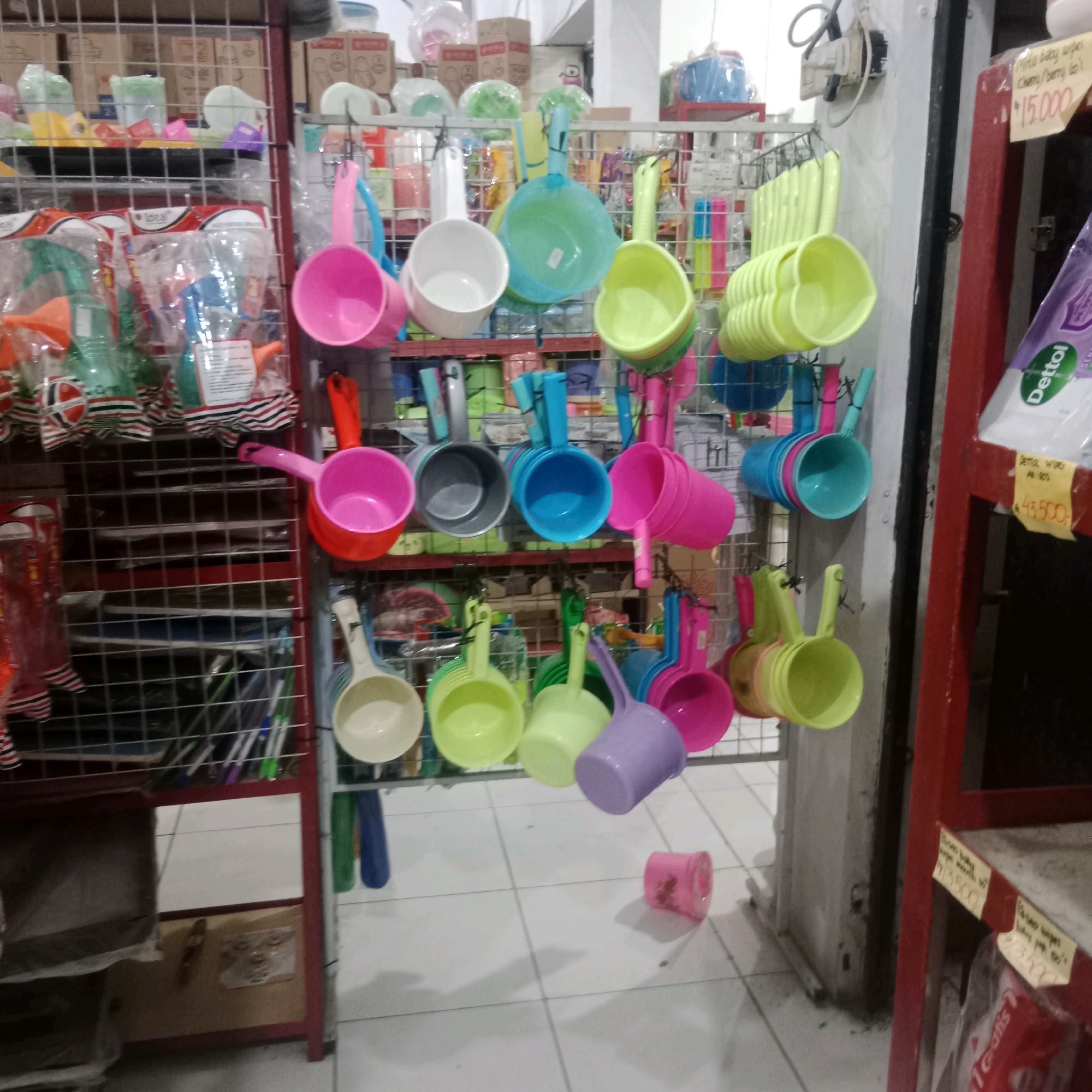 The image shows a store interior in Tegowanu Wetan, Indonesia, with a display of colorful plastic bath scoops hanging on metal racks. The scoops are in various bright colors, including pink, yellow, green, blue, and purple. Some are stacked together, while others are individually displayed. The racks are organized in rows and appear to be made of wire mesh. In the background, there are shelves stocked with other items, including boxed goods and plastic bottles. The floor is tiled and a red metal shelving unit is visible on the right side of the frame, displaying a price tag with Indonesian currency. A Dettol product is also visible on this shelving unit. The overall impression is that of a retail environment selling household or bath items.