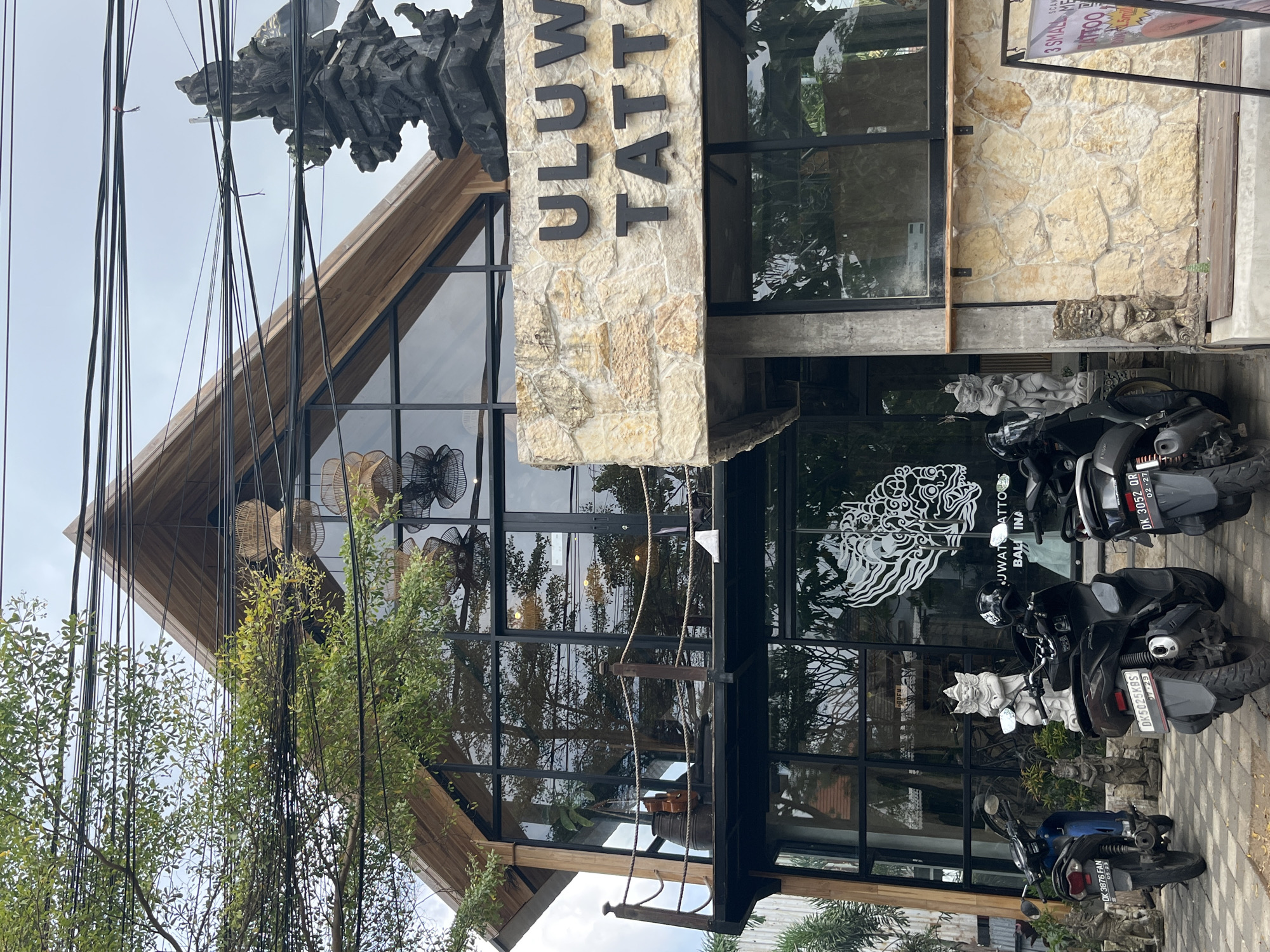 A wide shot captures the exterior of "Uluwatu Tattoo" in Kuta Selatan, Indonesia, a two-story building with a modern, angular wooden roof and large glass windows. The facade of the upper story is clad in rough-hewn beige stone, with black metal lettering spelling "ULUWATU TATTOO" across it. A large, ornate stone sculpture stands to the right of the building, above the entrance. The ground floor features floor-to-ceiling windows, revealing the interior and a prominent white tattoo design on the glass. In front of the building, three scooters are parked on a checkered paved area. Several small, grey stone statues of mythical creatures are strategically placed around the scooters and the entrance, some serving as decorative elements and others as bases for the scooters. A lush green tree with dense foliage partially obstructs the view of the building from the left side, its branches intertwined with a complex network of overhead electrical wires. The sky is overcast, suggesting a cloudy day.
