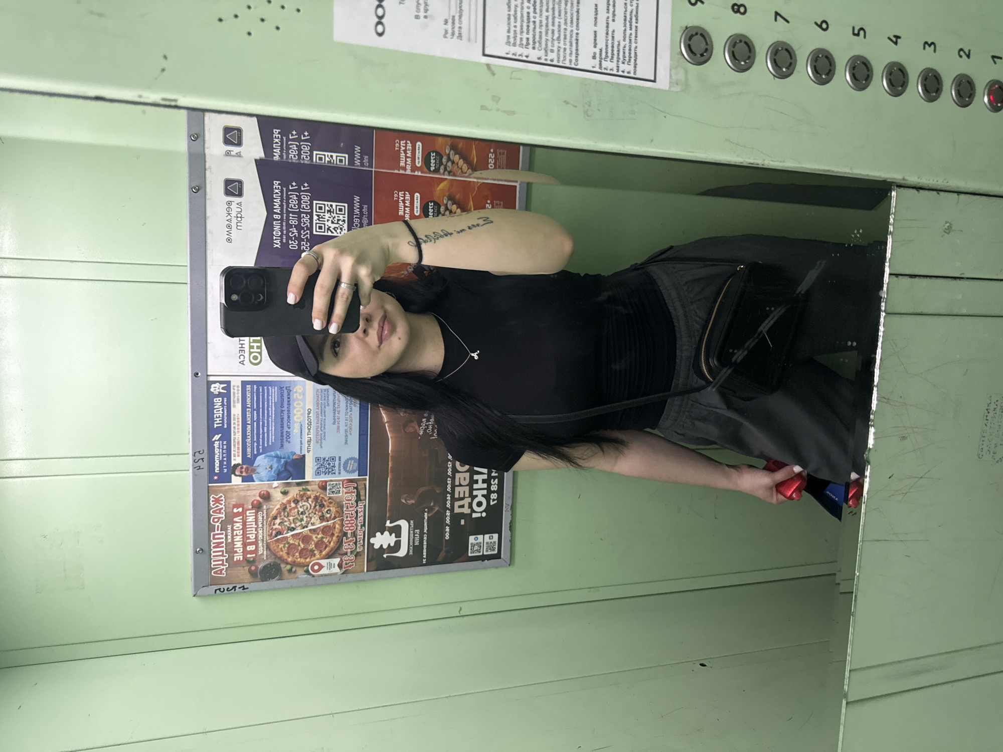 A young woman with long dark hair is taking a selfie in a mirror inside an elevator. She is wearing a black t-shirt, cap, and dark bottoms, and holding a black smartphone in her left hand. In her right hand, she is holding some red and blue items.
The elevator has light green walls and a mirrored surface, reflecting the woman and the advertisements posted on the elevator wall. These ads include advertisements for pizza, real estate services, and other businesses, with text in Russian. There are also elevator control buttons above the mirror.
Based on the text and information in the advertisements, the elevator is located in Mytishchi, Russia.