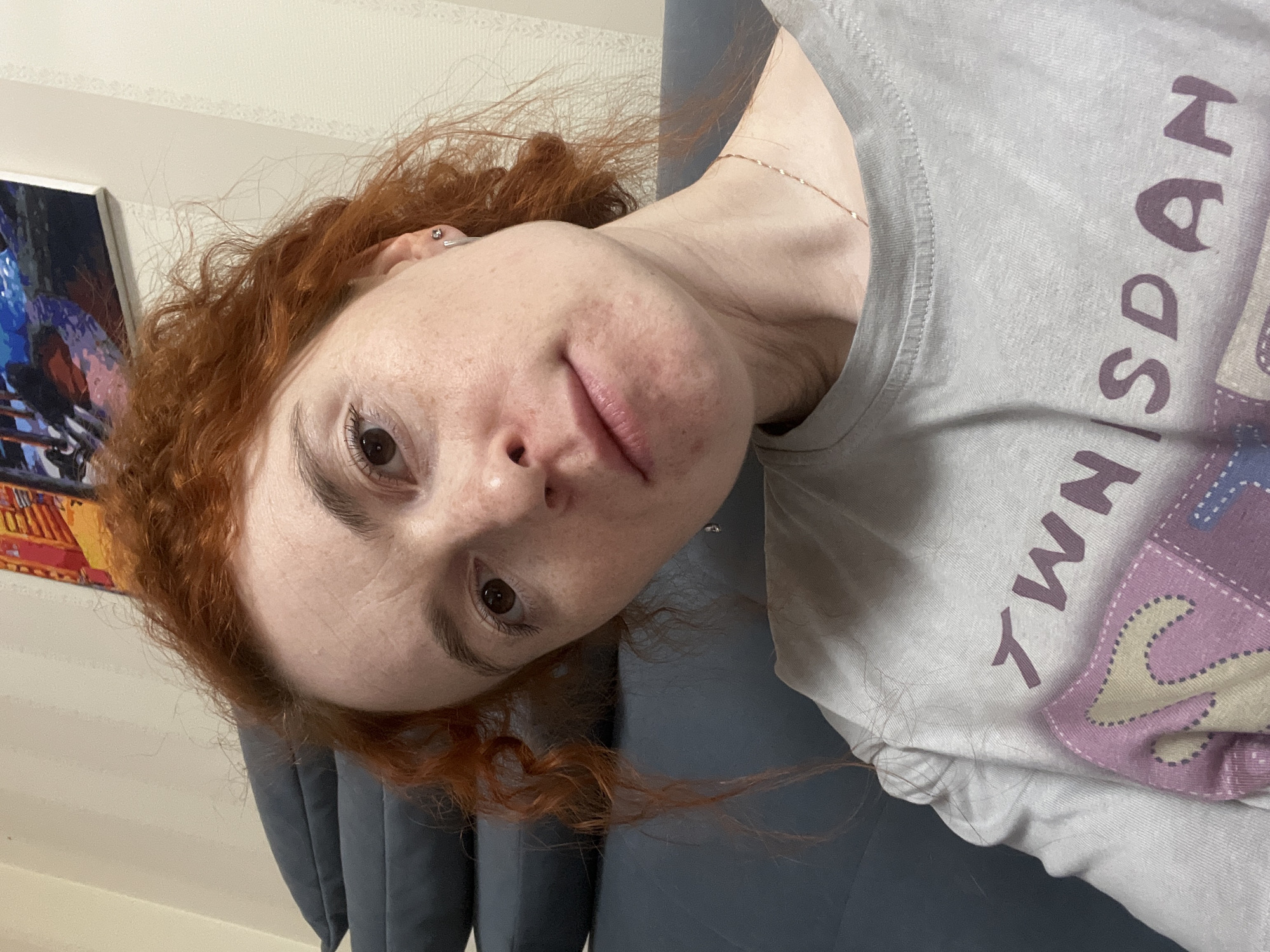 A fair-skinned woman with red curly hair is posing for a selfie. She is wearing a light grey t-shirt with purple lettering. A framed painting hangs on the wall behind her, and a blue upholstered chair is visible in the lower left corner of the frame. The specific location within Chaikovsky, Russia, is indiscernible from the available visual information.