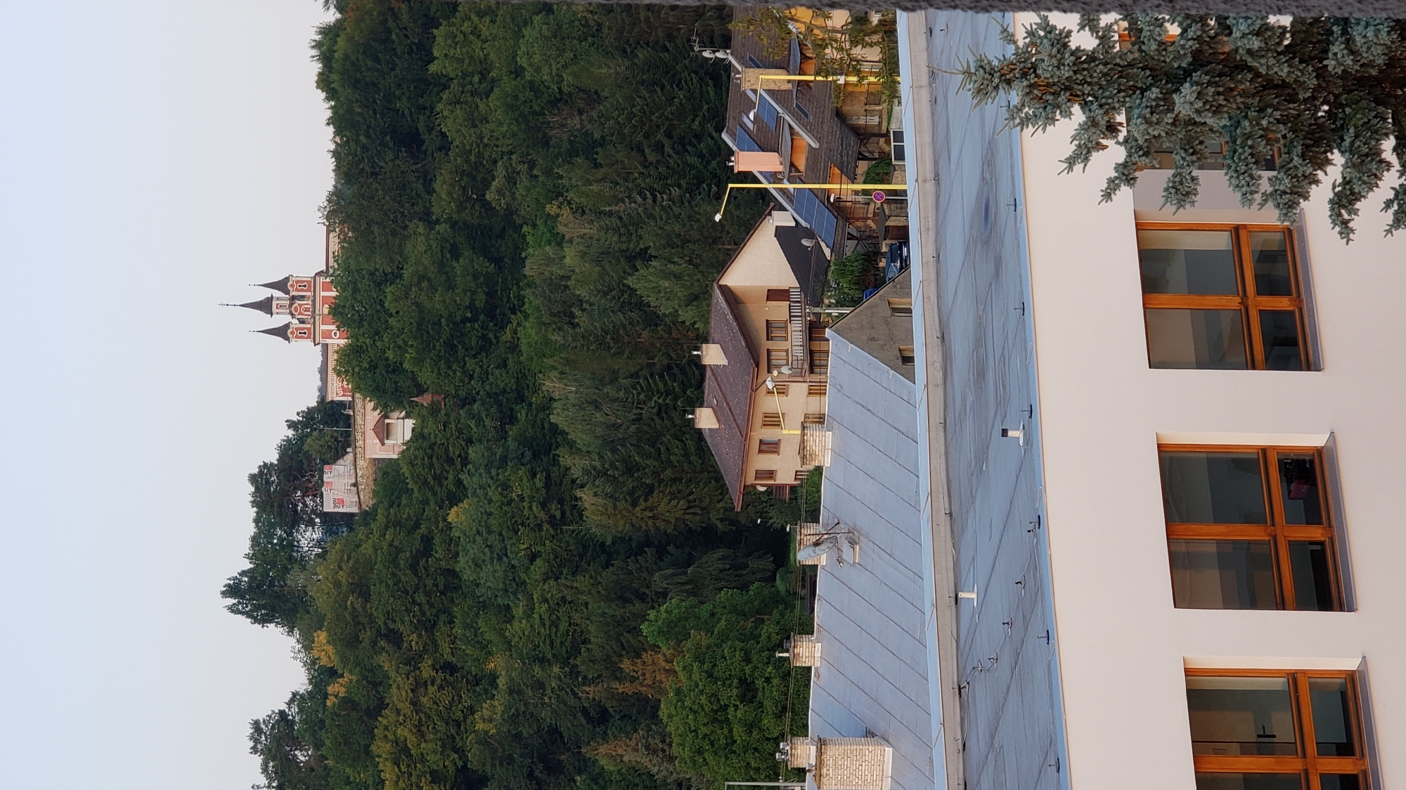 The image shows a cityscape with a prominent building on a hill in the background, set against a backdrop of lush green trees. The building on the hill features red and white architecture with multiple spires, resembling a church or a castle. In the middle ground, several houses with pitched roofs and chimneys are visible. The foreground is dominated by the flat, gray roof of a modern building with large, wooden-framed windows. A mature pine tree stands to the right of this building. The sky is clear and pale, indicating either early morning or late afternoon. The scene appears to be a view of the city of Prešov, Slovakia, from an elevated vantage point, possibly looking towards the historical center or a prominent landmark such as the Orthodox Church of the Holy Spirit or the Sigort church complex on a hill.