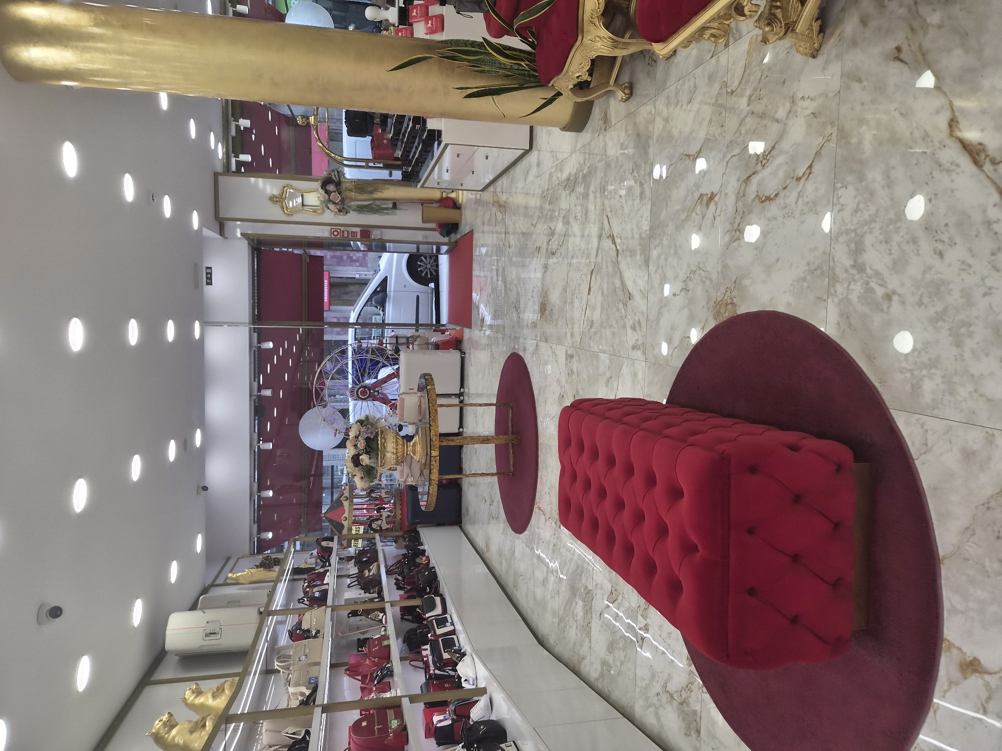 The image depicts the interior of a luxury retail store, likely a boutique specializing in handbags and accessories. The store is located on a street with a white van parked in front of the entrance, visible through the glass doors. The interior is bright and modern, with white marble-patterned flooring and white shelving units displaying an array of handbags in various colors, predominantly red, beige, and black. A prominent feature is a large, tufted red bench in the foreground, placed on a circular dark red rug. To the right, a tall, golden pillar stands out, and a gilded chair with red upholstery is partially visible. In the background, a golden table with a floral arrangement sits on a round rug. The ceiling features numerous recessed lights. The store's facade suggests it is located in a commercial area. While the city of Braga, Portugal, is mentioned, there are no specific visual cues in the image that confirm this location, such as signage or unique architectural features specific to Braga. No people are clearly visible in the image.