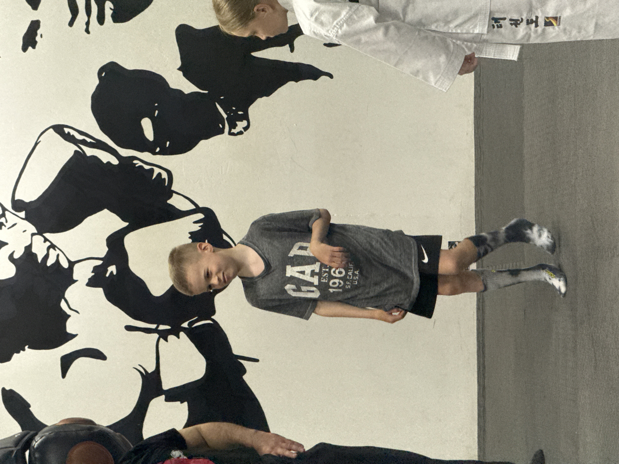 This is a medium shot, taken at an eye-level perspective, of two children standing on a dark grey mat in front of a white wall adorned with a large black graphic mural of stylized faces. The child on the left is a young boy wearing a grey GAP t-shirt with "EST. 1969 S.F. CALIF. U.S.A." printed on it, black shorts with a white Nike logo, and mismatched socks. He is looking slightly to his left with a contemplative expression. To the boy's left, partially out of frame, is an adult wearing dark clothing and a black helmet with large, round ear coverings. The child on the right is a young girl wearing a white taekwondo uniform with black Korean text and a yellow logo on the sleeve. She is facing away from the camera, looking to her left, and her hands are clasped in front of her. The overall scene appears to be a martial arts studio or a similar training facility. There is no information in the image to suggest the location is Moscow, Russia.