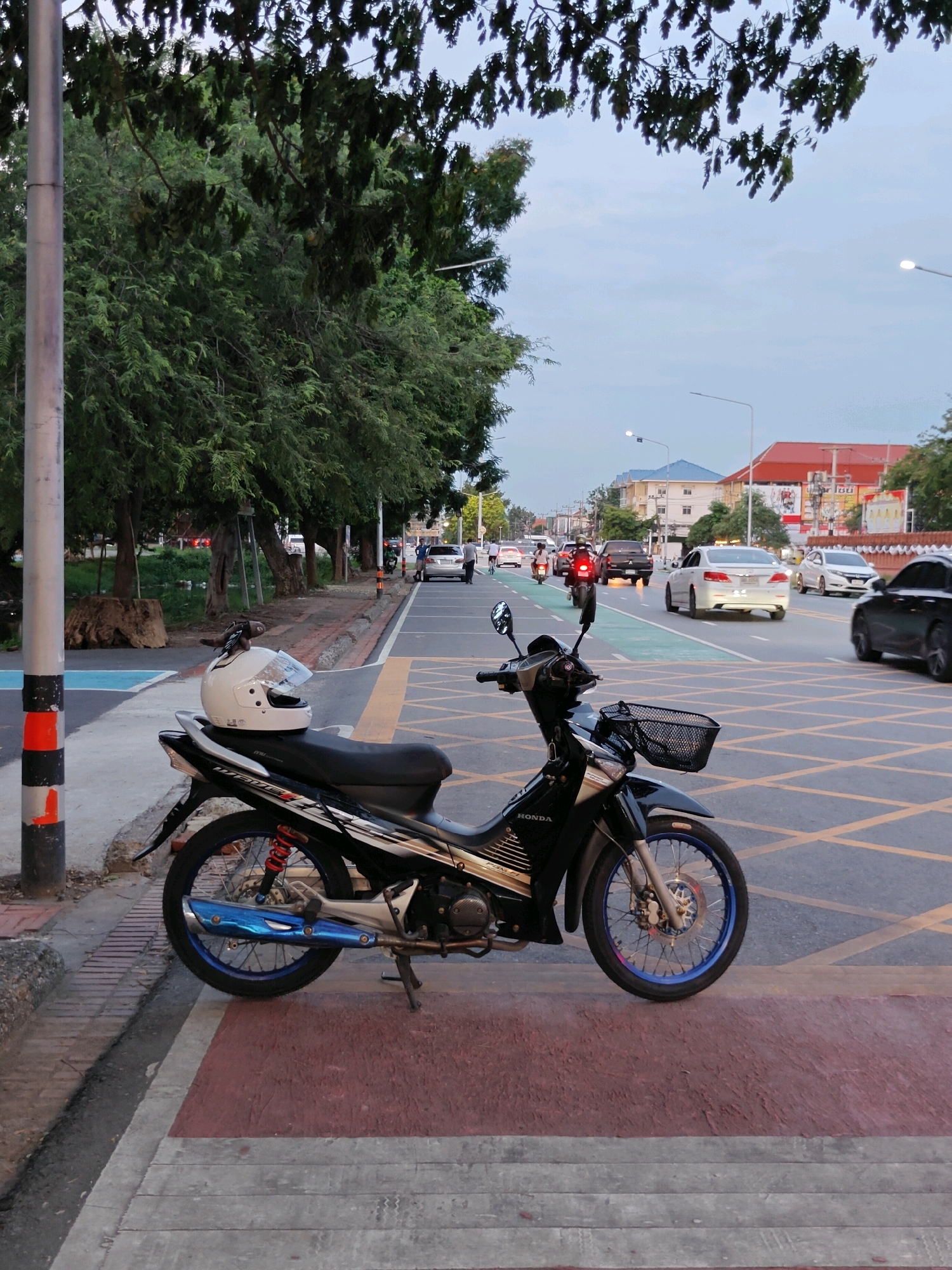The image shows a black and silver Honda motorcycle parked on a pedestrian walkway in Phra Nakhon Si Ayutthaya City Municipality, Thailand. A white helmet with a clear visor is placed on the motorcycle's seat. The walkway has a textured, reddish-brown surface with grey and white striped sections. Behind the motorcycle, a street is visible with several cars and a few people. The street has multiple lanes with yellow and white markings. Trees line the left side of the street, and buildings with red and blue roofs are visible in the background. The sky is overcast and appears to be late afternoon or early evening. The overall scene depicts a typical urban streetscape in Thailand.