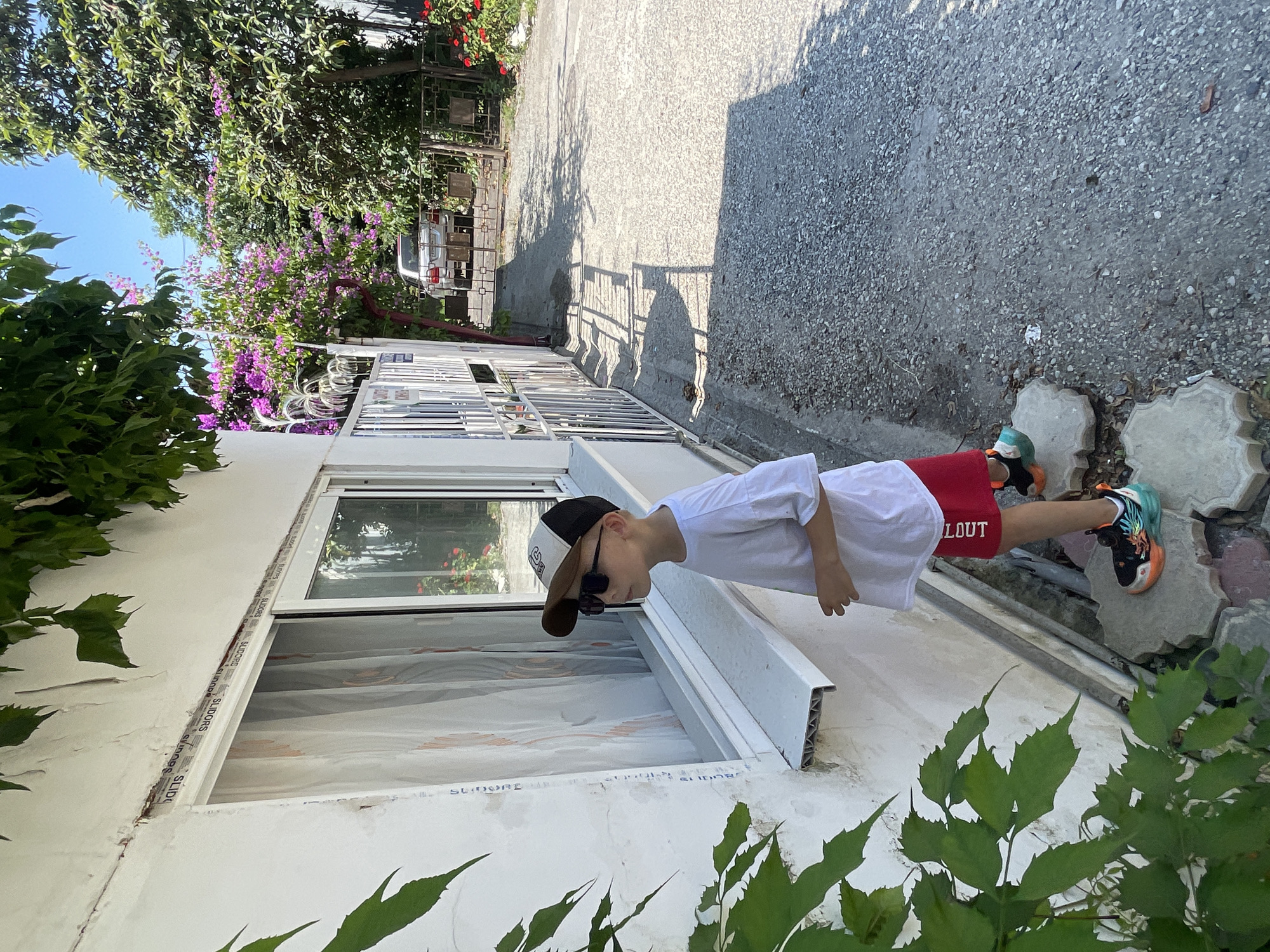 In Gagra, Abkhazia, a young Caucasian boy stands outside a white building on a sunny day. He is wearing a white t-shirt, red shorts with white lettering, a baseball cap with a brown brim, and sunglasses. His shoes are multi-colored with black, orange, and green accents. The boy is looking towards a window with sheer white curtains. Green ivy grows on the left side of the building, and purple bougainvillea flowers bloom in the background to the right. A gravel path runs alongside the building, and a car is visible through a gate in the distance. The scene suggests a residential or commercial setting in a warm climate.