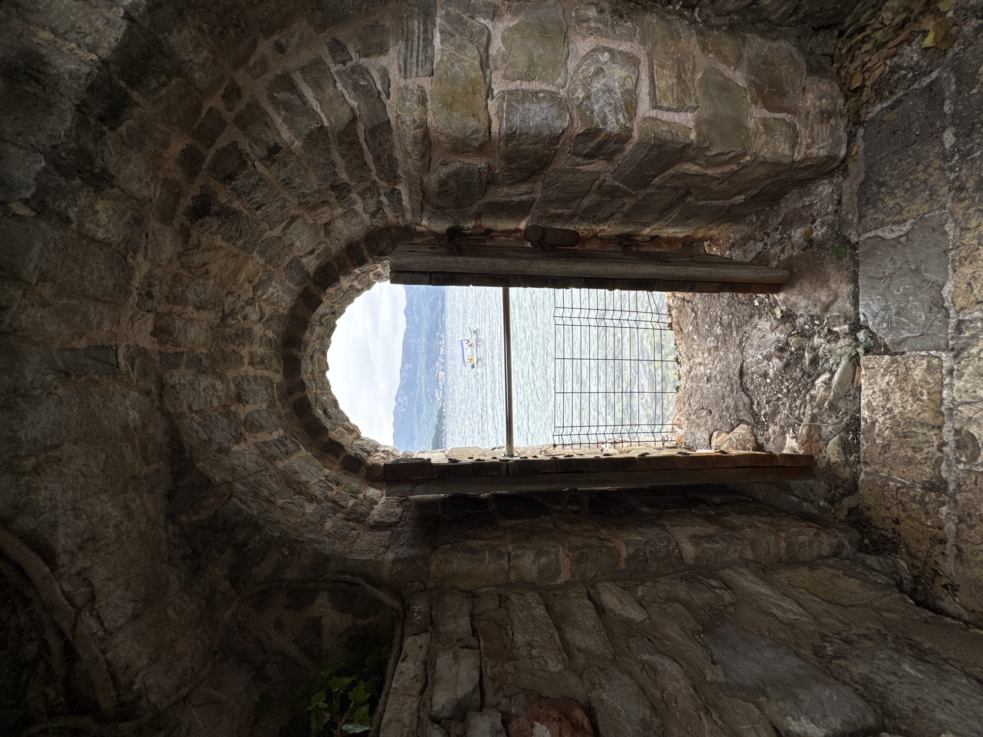 The image shows a stone window looking out at a lake. The walls of the window are constructed of irregularly shaped stones. A partial metal fence is visible in the middle ground, and a blue lake stretches to the horizon, with a blurry mountain range in the background. A boat can be seen on the lake. The image appears to be taken from inside a stone structure in Budva, Montenegro.